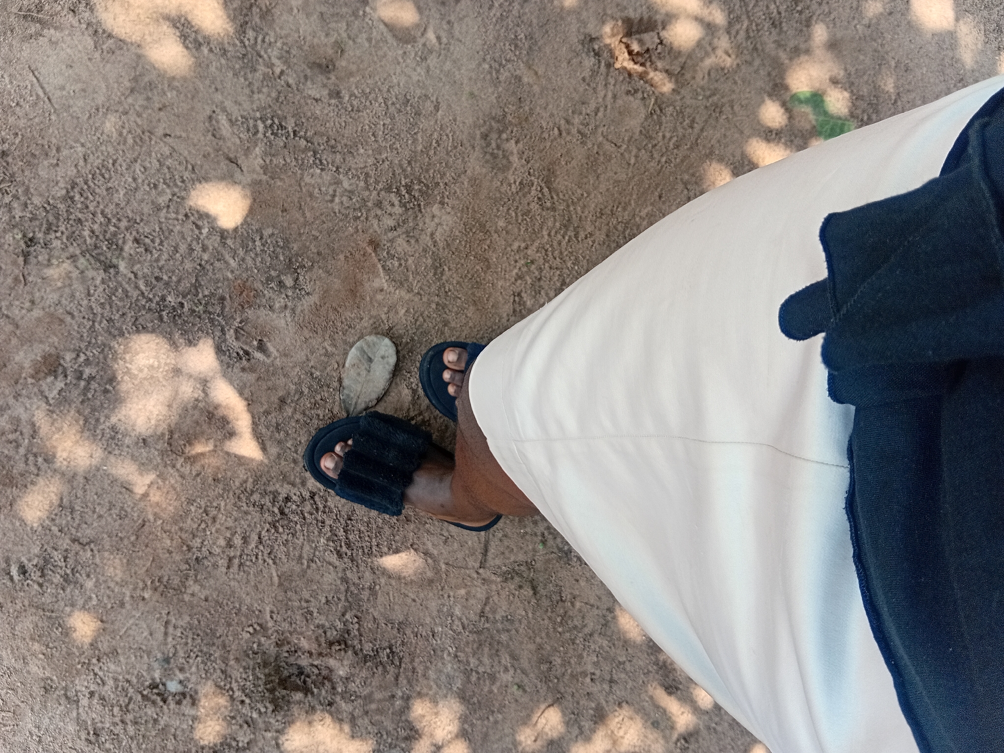 This is a high-angle, outdoor shot taken in Ejule, Nigeria, focusing on the lower half of a person's body. The ground is composed of sandy soil with scattered leaves and light patches of sunlight creating dappled shadows. The person is wearing a light-colored garment, possibly trousers or a skirt, and dark blue, fluffy slippers. The overall scene suggests a casual outdoor setting in Nigeria.