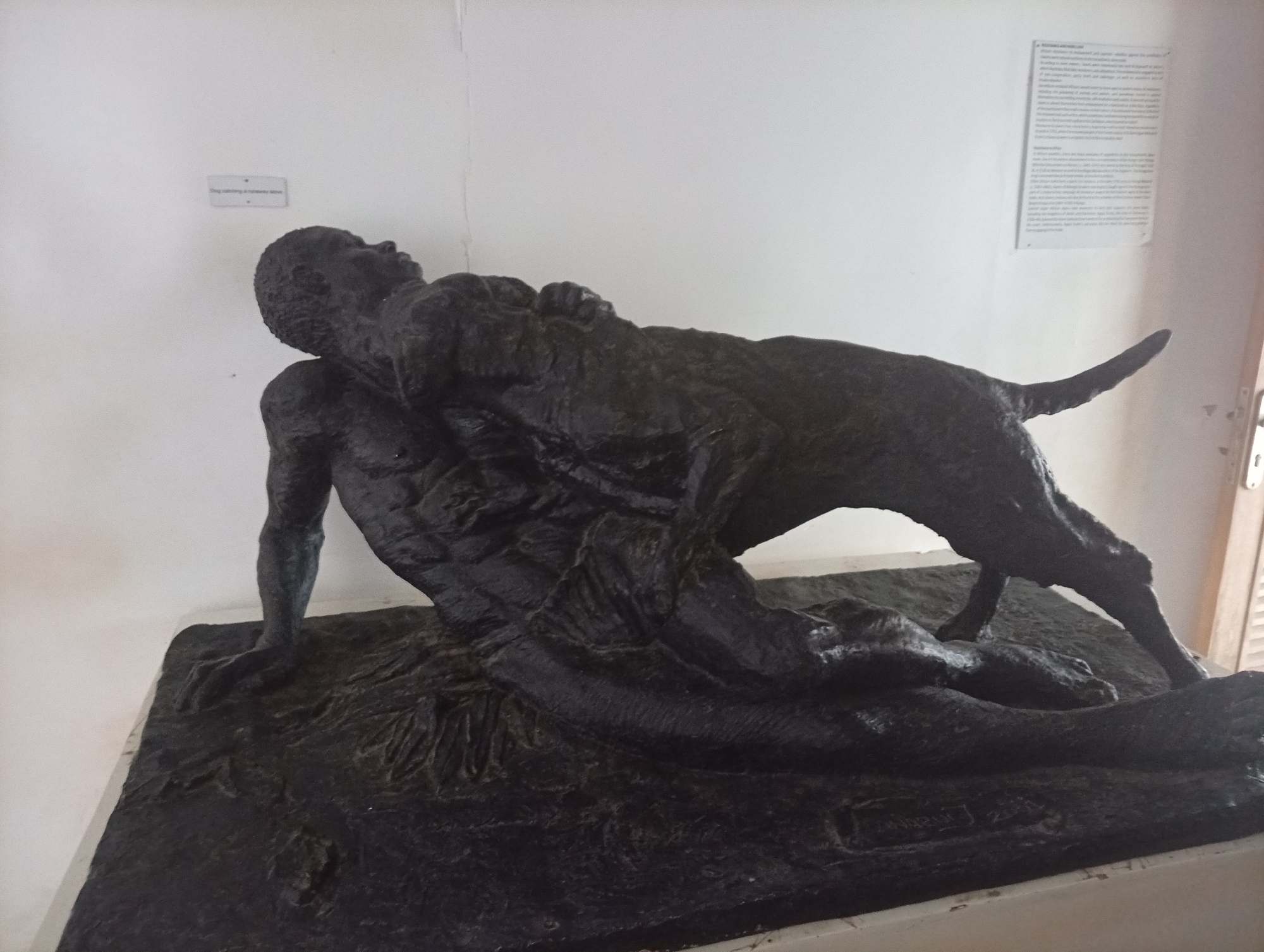 The image shows a dark bronze sculpture of a man and a dog, located indoors in Badagry, Nigeria. The sculpture depicts a man lying on his back with a dog standing over him. Both figures have textured surfaces. The scene is set on a rectangular base. A sign is visible on the wall above the sculpture, and another sign containing printed text is above the sculpture to the right. The room has white walls.