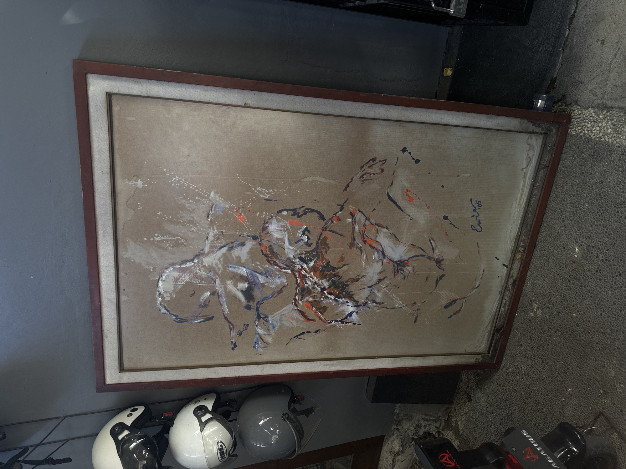 The image shows a framed abstract painting leaning against a wall in an indoor setting. The painting, predominantly brown, features dynamic brushstrokes in white, orange, and blue. A signature, seemingly "Euri's '05", is visible in the upper right corner. Below the painting, three motorcycle helmets are arranged: two white with black accents and one grey. Part of a black structure is visible above the painting. The floor adjacent to the painting consists of small, grey pebbles. A black object with red detailing is partially visible in the lower right corner. The image was taken in North Kuta, Indonesia.