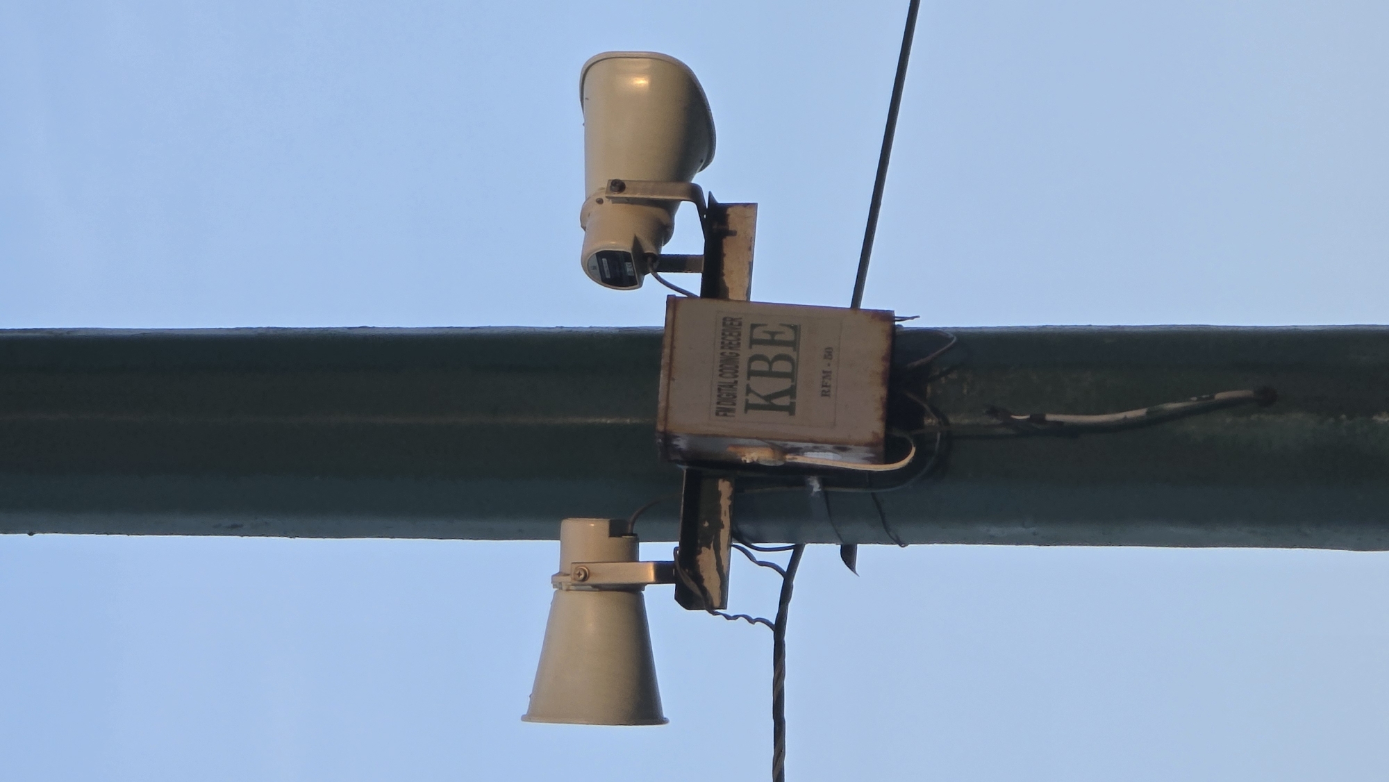 The image depicts two outdoor loudspeakers, one facing upwards and the other downwards, mounted on a horizontal, dark green I-beam against a light blue sky. Between the speakers is a metal box labeled "FM Digital Coding Receiver KBE RFM-50." Electrical wiring is visible, connecting the components and running along the beam. The scene is devoid of people. The location is specified as Nha Trang, Vietnam, though no local landmarks or details are visible to confirm this.