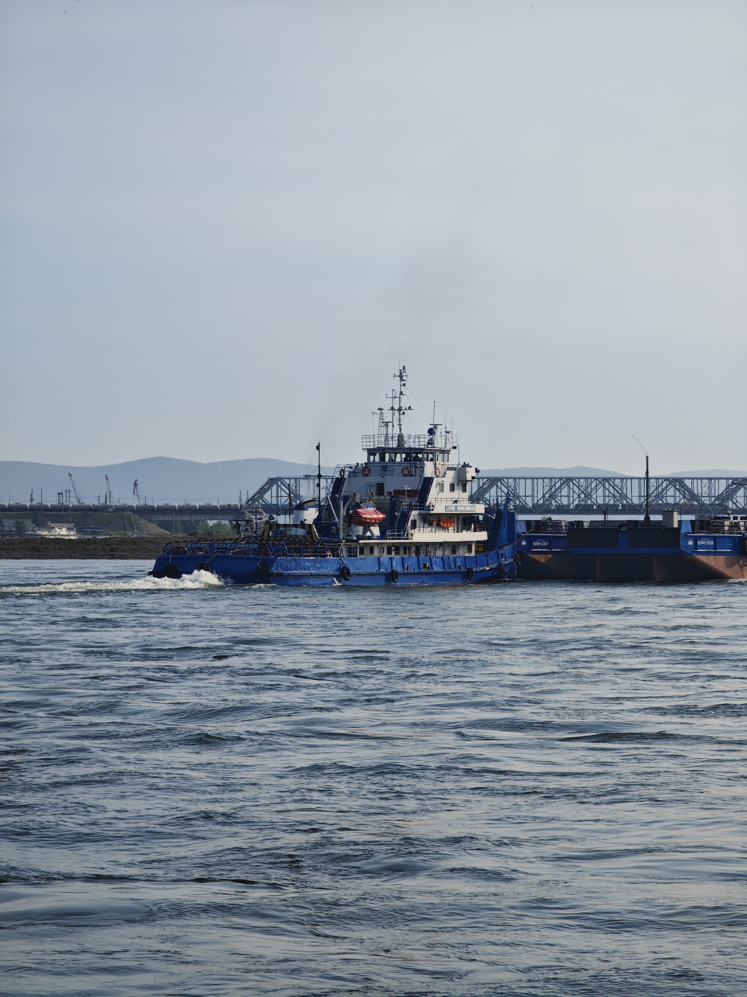A large blue and white tugboat navigates a wide, rippling river. The tugboat, named "БОРИС КОЛЕСНИКОВ" (Boris Kolesnikov), is pushing a large barge. The barge has "БПН-2510" (BPN-2510) and "БПН-2508" (BPN-2508) written on it. In the background, a large, trussed bridge spans the river. Beyond the bridge, cranes are visible, suggesting ongoing construction or industrial activity on the riverbank. Rolling hills form the distant horizon under a hazy, overcast sky. The scene appears to be set during the daytime, with soft, diffused light. The river is choppy, indicating a moderate current or wind. The overall atmosphere is one of industrial transit and transportation, with no visible people or specific emotional cues. The location is identified as Krasnoyarsk, Russia, which aligns with the presence of a large river and industrial infrastructure.