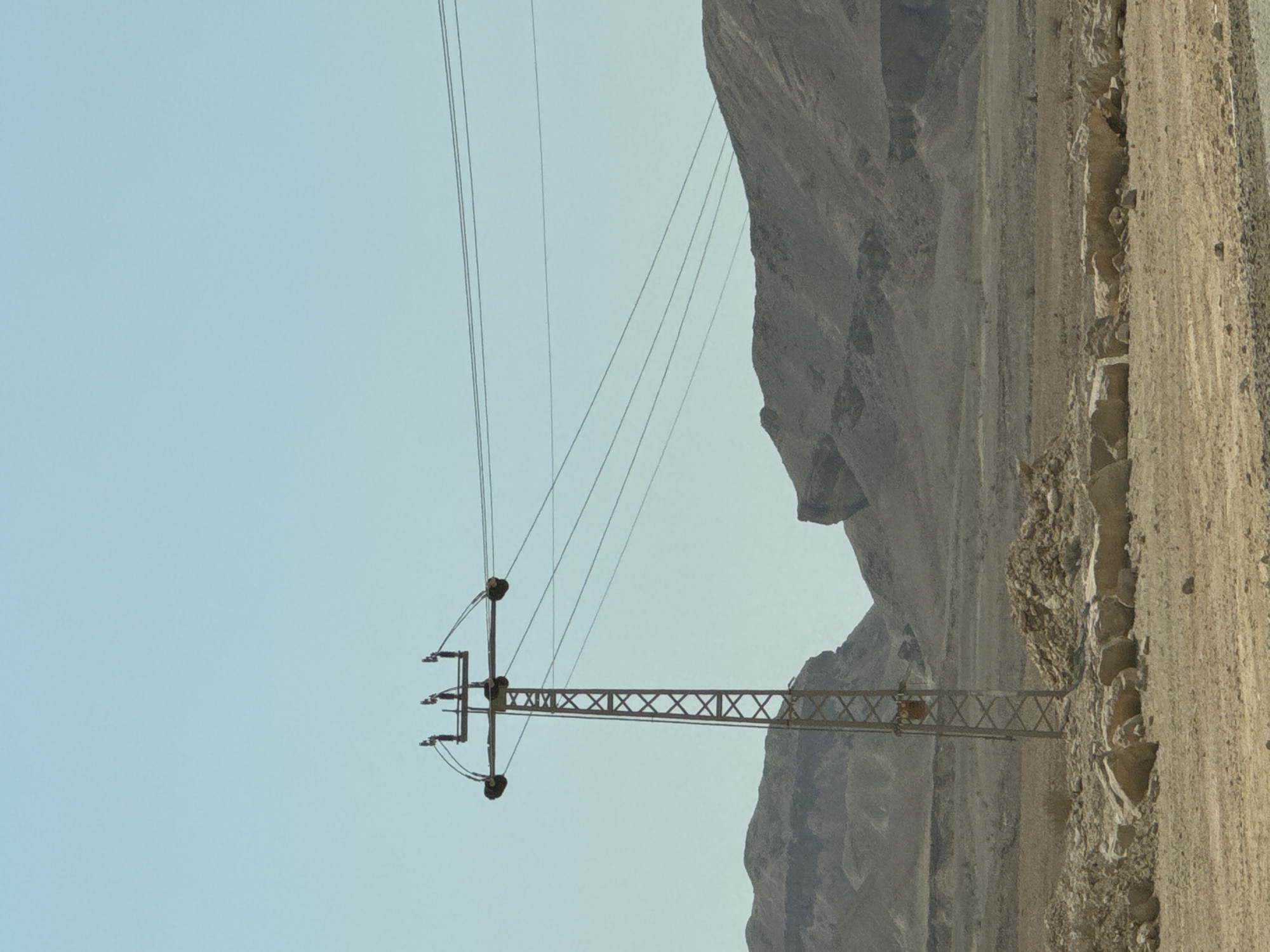 A solitary, tall, lattice-style electrical pylon stands in a desert landscape under a clear, pale blue sky. The pylon is made of metal and has a lattice structure with visible cross-bracing. It supports multiple electrical wires that stretch horizontally across the frame, disappearing into the distance. In the foreground, the ground is dry, sandy, and littered with rocks and stones. The middle ground features a sparse, arid terrain with low-lying vegetation. In the background, rugged, barren mountains dominate the horizon. The scene depicts a remote, undeveloped area within the Ramat Negev Regional Council in Israel, characterized by its harsh, dry environment.