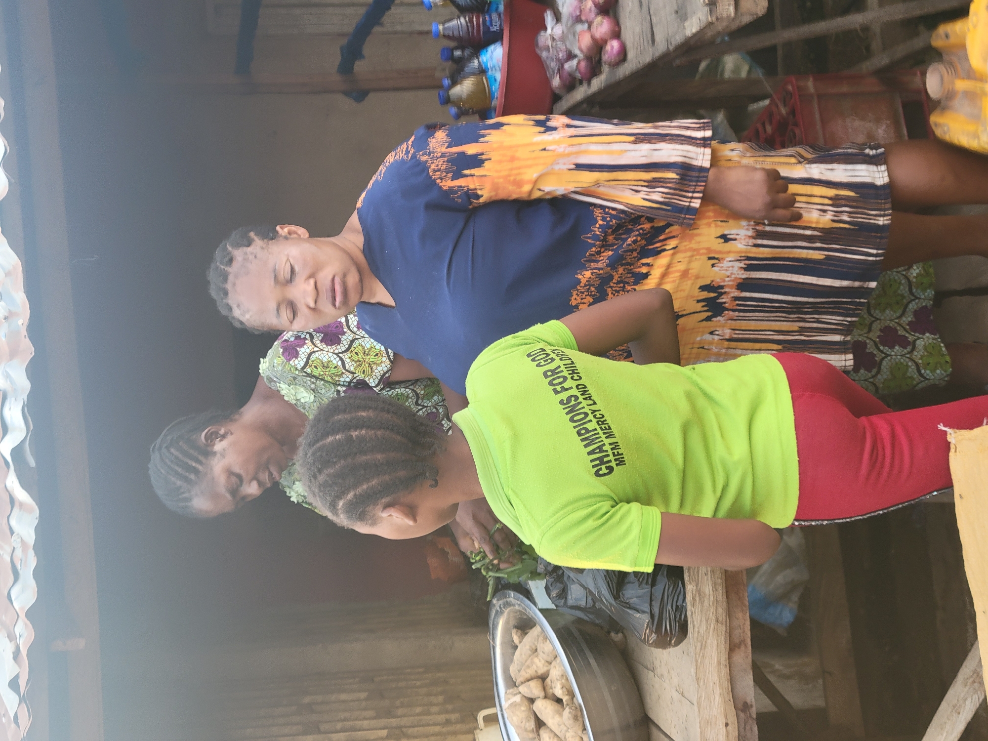 In a moderately lit outdoor market setting in Madalla, Nigeria, three females engage in preparing food items. The backdrop showcases items such as bottled drinks and red onions.
The first female, dressed in a blue and patterned yellow dress, stands prominently. The second female, dressed in a green and purple patterned top, is seen near a child in a bright green t-shirt with red pants. The child’s shirt has “Champions for God” printed on it. They stand near a wooden table where food preparation is taking place. A black plastic bag and a metal bowl containing several food items are present on the table.