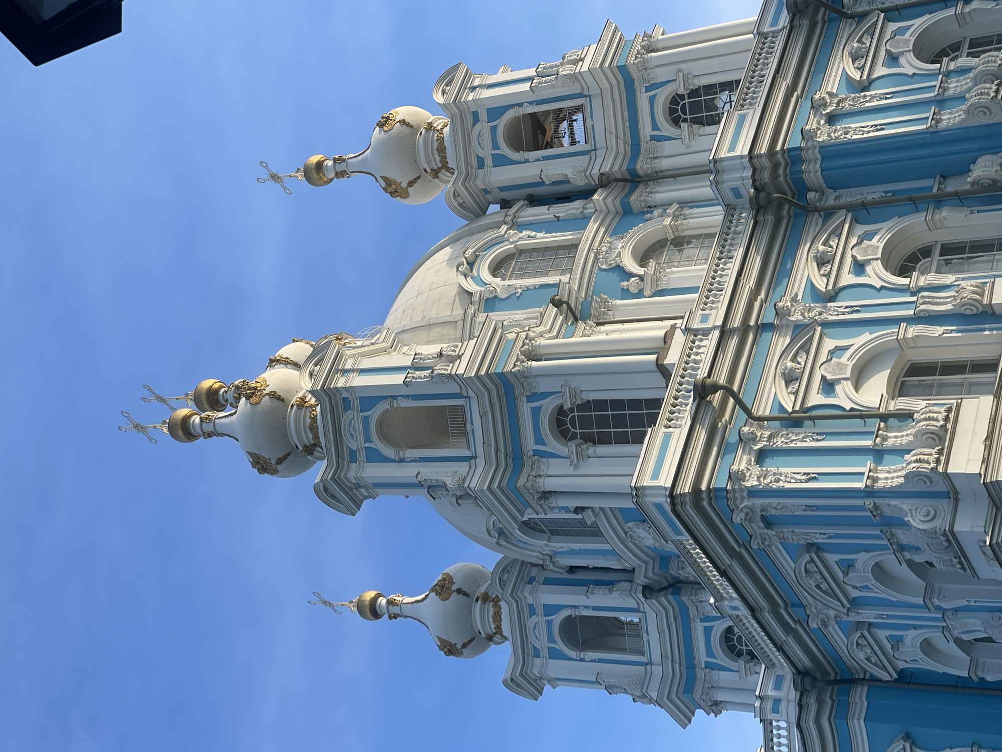 The image displays a low-angle exterior view of a large, ornate cathedral or church, set against a clear, bright blue sky. The building exhibits a striking architectural style characterized by predominantly sky-blue and white facades, accented with intricate white stucco ornamentation and gold details. Multiple white, bulbous domes, some with a distinct onion shape, rise above the structure, each capped with a golden sphere and an Orthodox cross. At least five such crosses are visible across the upper sections of the building. The central dome is larger and white.
The facade features numerous arched windows, some with internal grilles, and is adorned with classical white columns, elaborate balustrades on various levels, and highly detailed rococo-style stucco elements, including volutes, scrolls, garlands, and carved facial motifs positioned above arches and along cornices. Several towers incorporate open archways, indicative of belfries. Visible elements of the building's infrastructure include dark-colored downspouts running vertically along the blue sections. The clear blue sky suggests fair weather conditions and ample natural light illuminates the structure, highlighting its details and casting subtle shadows. No individuals or other significant objects are present in the scene. The location is in the city of Unknown, Russia.