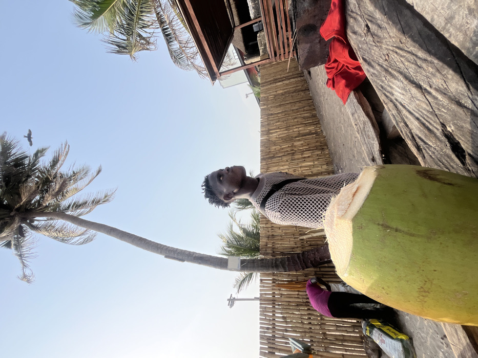 A young person looks up at a bird flying past a palm tree against a bright sky. The person is wearing a mesh shirt with black straps, stands next to a large, green coconut that has been recently chopped open. The background features a bamboo fence, a weathered wooden surface, and a partially visible building structure. Some clothing items, possibly a red shirt and purple leggings, are also present in the frame. The photo seems to have been taken at an angle, slightly tilted upwards.