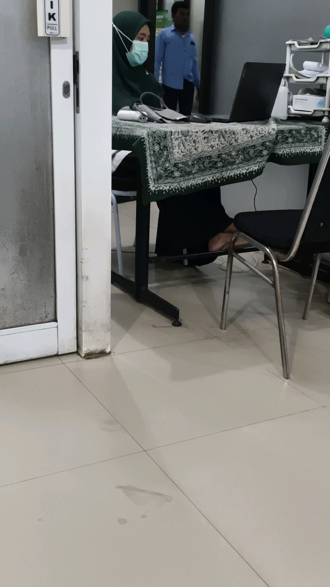 A veiled female health professional, wearing a teal face mask and a dark green hijab, sits behind a table covered with a green and white patterned cloth in what appears to be a clinic or hospital setting in Rejoagung, Indonesia. On the table are medical instruments, including a blood pressure cuff and a digital thermometer, along with a laptop. To the right of the table, a metal-framed chair with a black seat is occupied by unseen legs and feet. A second individual, a male wearing a blue shirt, stands in the background. The floor is tiled with a light-colored material. A sign with the letters "IK" and "PULL" is visible on a white door frame to the left of the frame.