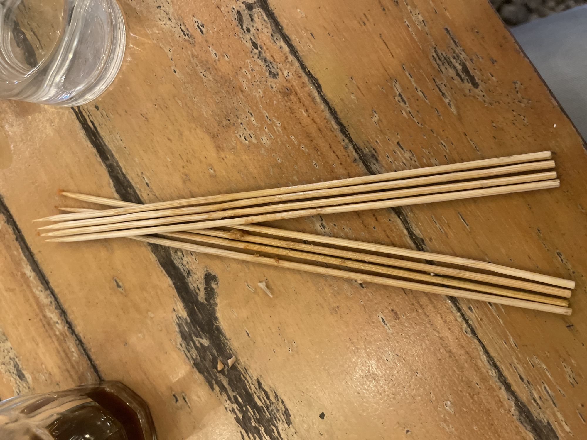 The image shows a close-up view of several wooden skewers lying on a wooden table. The skewers are arranged in a fan-like pattern, with their pointed ends facing towards the upper left of the frame. Some of the skewers appear to have remnants of food on them. To the upper left of the skewers, a portion of a clear glass, possibly a drinking glass, is visible. The table surface is made of wood with a textured grain and dark, irregular markings. In the bottom left corner, a dark brown liquid in a glass container is partially visible. The scene appears to be indoors, possibly in a restaurant or dining setting. The lighting suggests it could be daytime or that artificial lighting is being used. There is no visible text in the image.
