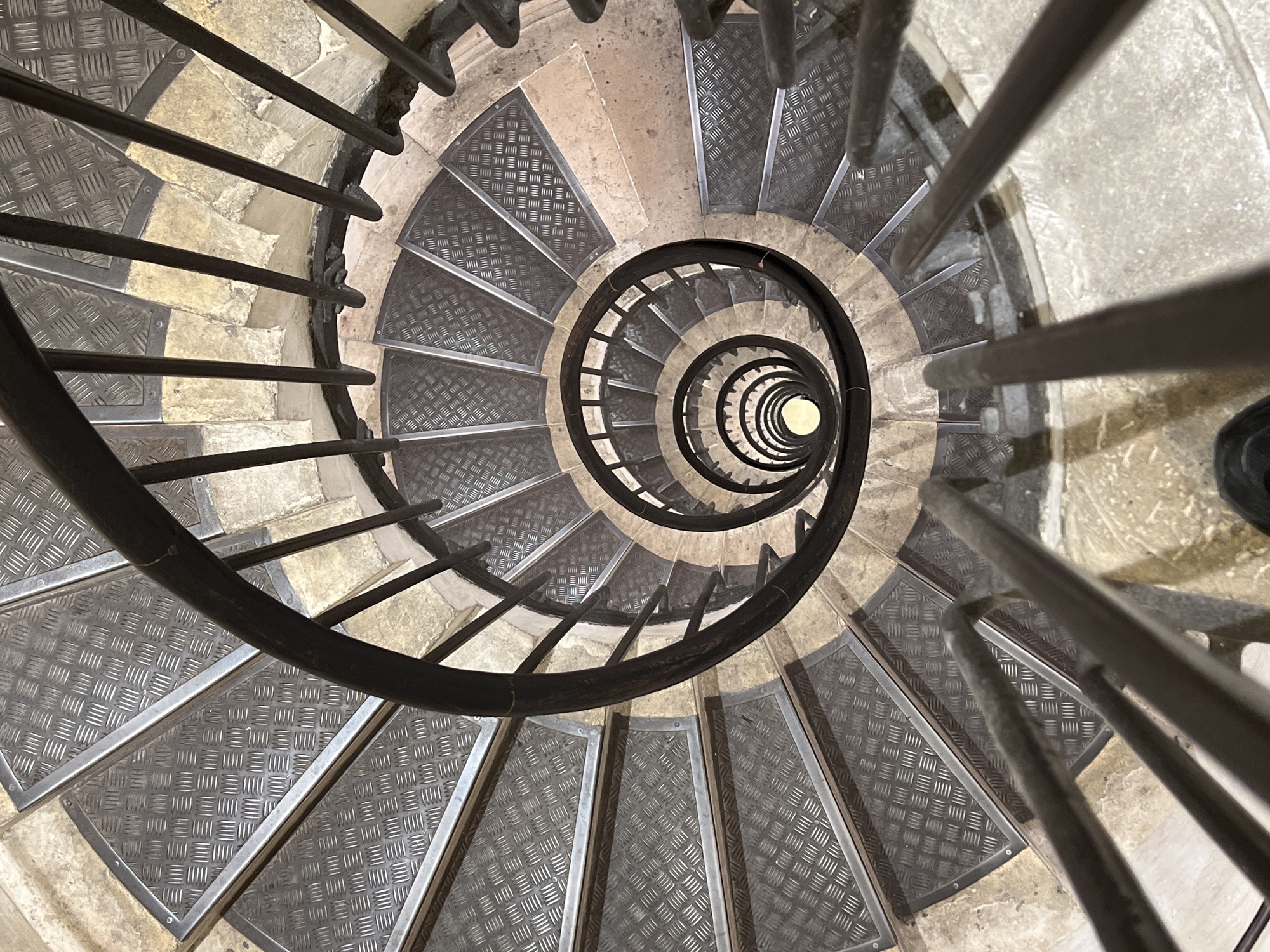 This top-down, high-angle shot presents a dizzying view of a spiral staircase, looking directly down its central core. The staircase descends into a bright, circular light source at its deepest point, suggesting a light well or an opening to a lower level.
The steps are a combination of light-colored, worn stone or concrete and dark grey metal plates. Each metal tread features a distinct raised diamond plate (checker plate) pattern, designed for anti-slip functionality, and is framed by a thin metal border. These metal inserts contrast with the older, rough-textured stone structure of the steps and risers.
A robust, dark-colored handrail or balustrade, possibly made of wrought iron, spirals downwards along the outer edge of the steps. It consists of multiple parallel bars, reinforcing the helical form and creating a sense of enclosure. The repetitive pattern of the metal steps and the curving railings emphasize the depth and architectural rhythm of the stairwell.
No individuals are present, and no specific event or activity is depicted. The scene is an architectural study focused on the structure and materials of the staircase. The lighting is concentrated at the bottom of the well, illuminating the descent. There is no visible text. This functional and visually striking staircase is located in Paris, France, its blend of historic stone and modern metal suggesting its use in a public building or tower.