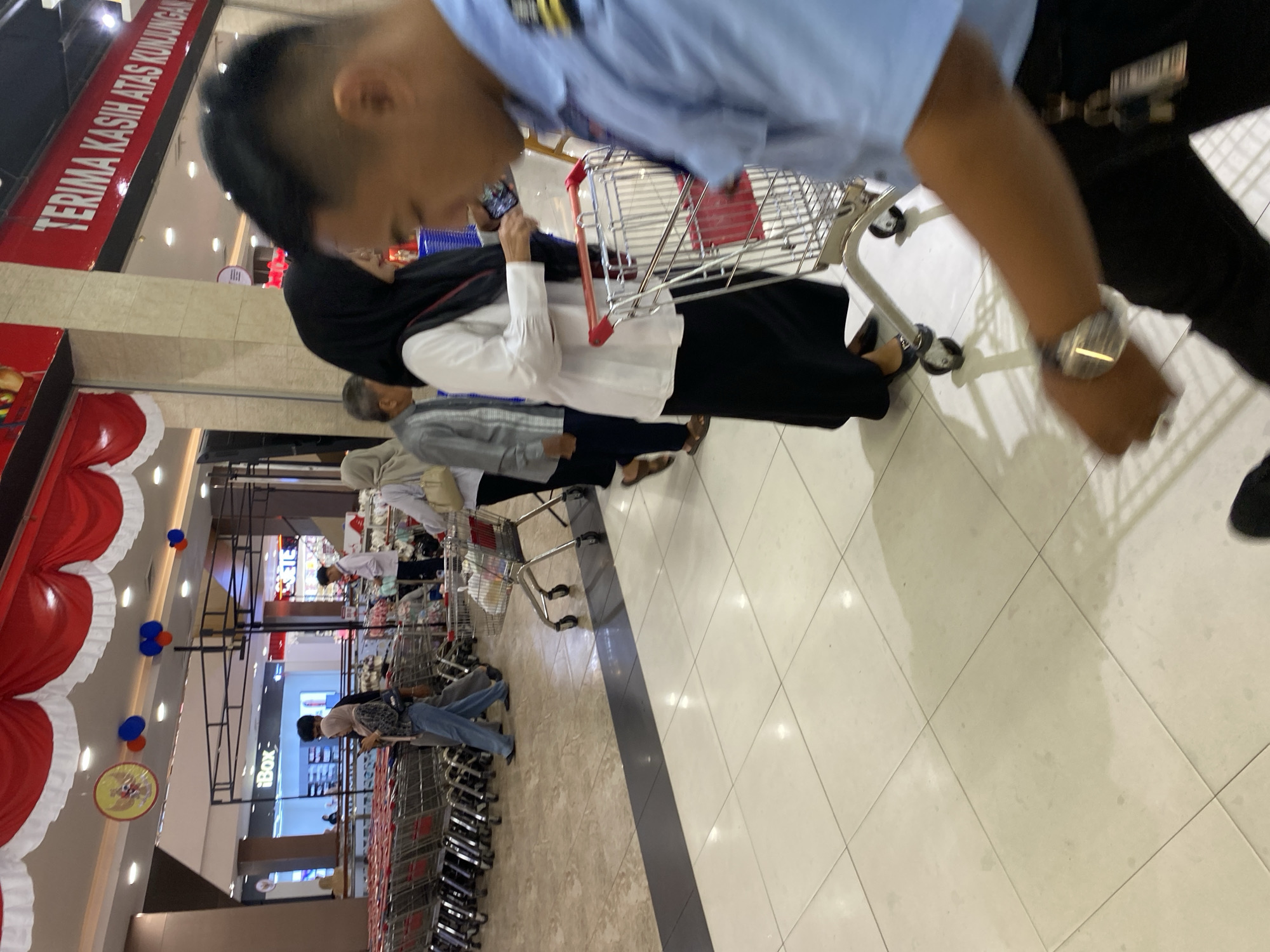 The image depicts a scene inside a shopping mall, likely in Banda Aceh, Indonesia. In the foreground, a person wearing a light blue uniform and dark pants is partially visible, their arm and wrist with a watch are in focus, suggesting they may be a security guard or mall staff. They appear to be walking or standing. Behind them, several people are walking or browsing. A woman in a black hijab and white shirt, and a long black skirt, is prominent, holding a mobile phone. Another person in a grey garment and headscarf is visible to her left. Further in the background, more shoppers and numerous empty shopping carts are arranged in rows, indicating a commercial environment. The mall features a tiled floor, overhead lighting, and decorative elements like red and white banners with Indonesian flags and circular emblems. A prominent red sign with white text, "TERIMA KASIH ATAS KUNJUNGAN" (Thank you for your visit), is visible on the right side of the frame, common in Indonesian businesses. The overall atmosphere suggests a busy, well-lit indoor retail space.