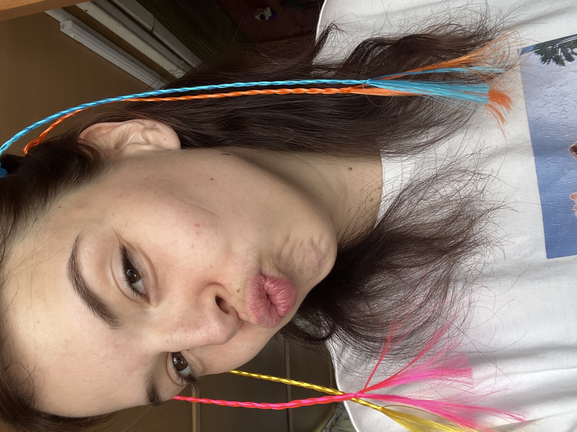 A young adult female is depicted in a close-up, selfie-style portrait with an intentionally pursed-lip expression and a slightly squinted or winking left eye. Her dark brown hair is parted on the left and worn down, with multicolored hair strands or extensions braided into her natural hair. On the left side of her head, yellow and pink braided strands with feathered ends are visible. On the right side, blue and orange braided strands, also with feathered ends, are present. She has a fair complexion with minor skin irregularities. She is wearing a white short-sleeved t-shirt, on the front of which a partial graphic print of a blue sky, clouds, and the top of a palm tree is visible. The background is an indistinct indoor setting with warm brown walls and faint vertical and horizontal elements that may be shelving or window blinds. A dark, skull-like object is vaguely discernible on a surface in the upper right background. The scene is located in Мшинская, Russia.