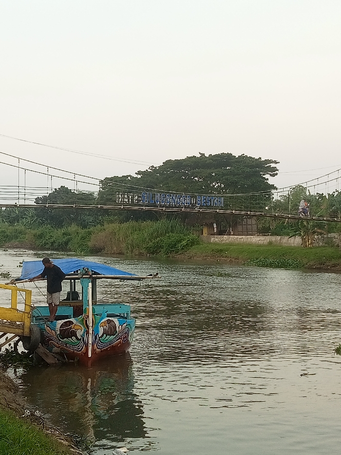 The image shows a brightly colored boat docked along the bank of a river in Jakenan, Indonesia. The boat is blue, yellow, red, and green and features a painted design of birds. A man is standing on the deck of the boat. The boat is partially covered with a blue tarp. A wooden bridge is in the background with a sign that reads, "GATE 2 SILUCONGOO BESTARI." The river has still, murky water. There is dense green foliage surrounding the river. The sky is clear and light grey. The coordinates provided locate the scene near the town of Jakenan, Indonesia, which is situated in the Pati Regency region of Central Java province.
