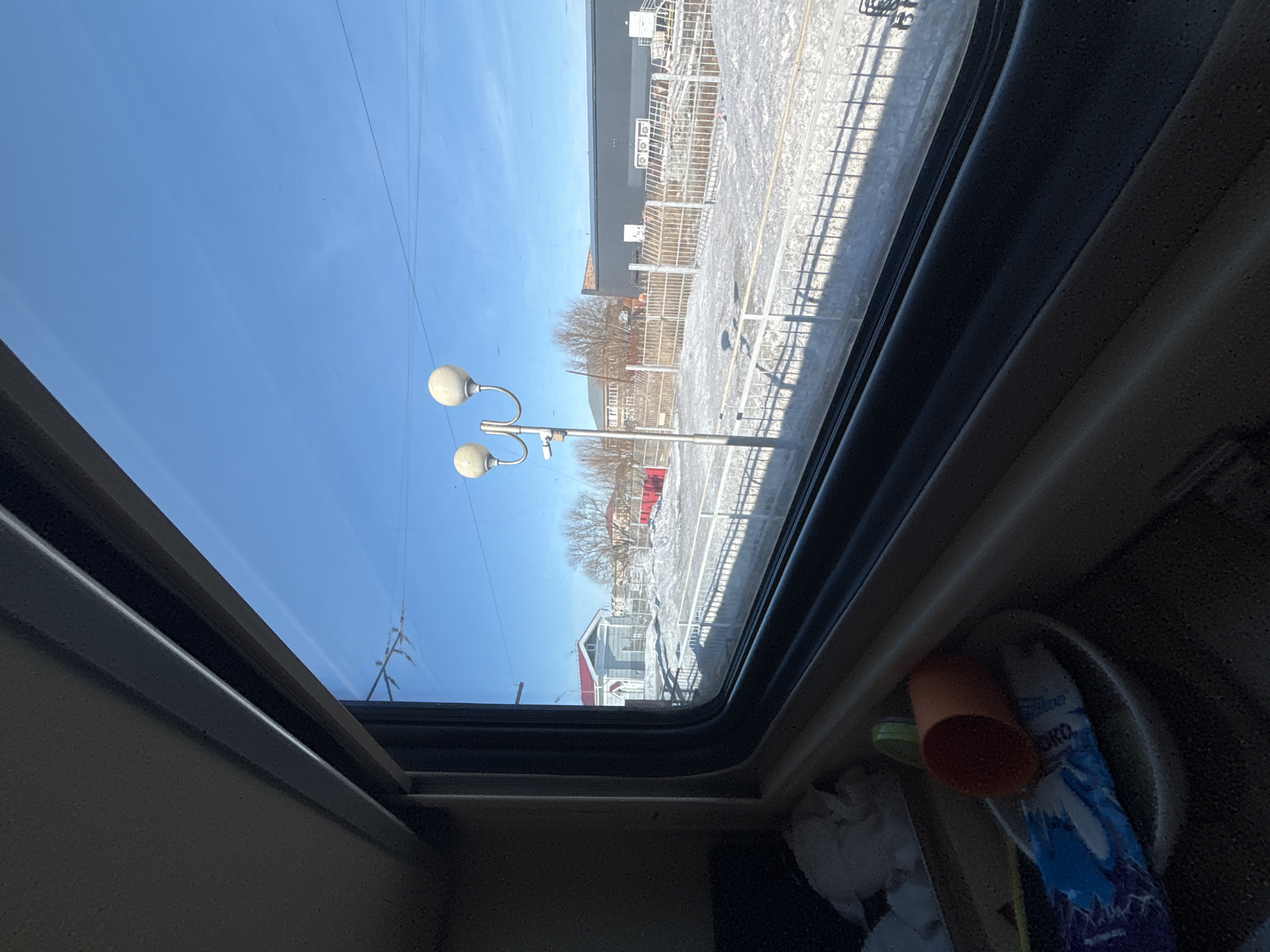 The image captures an exterior view through a window, likely from a train, in the city of Chany, Russia. The foreground shows a dark, textured interior surface with various objects: an orange cylindrical cup lying on its side, a white bowl, a blue and white wrapper (partially visible, displaying "ОКО"), a green cap, and crumpled white paper.
Beyond the window, a sunny, clear blue sky dominates the upper portion of the frame. The ground is characterized by patches of snow and bare dirt, indicating a cold season. Train tracks are visible, partially covered by snow, running parallel to a light-colored metal fence that extends across the mid-ground. A prominent silver lamppost with two spherical, opaque white light fixtures and a small dark camera attached to its pole stands left of center.
In the background, on the left, a small light-grey building with a red pitched roof is visible. To the right, a large, dark grey industrial-style building with a flat roof and several white rooftop ventilation units is present. Further back, a multi-story building with multiple windows can be seen, partially obscured by several bare deciduous trees. Thin dark power lines traverse the clear sky. No individuals are visible in the scene. The setting appears to be a train station or railway area in Chany, Russia.