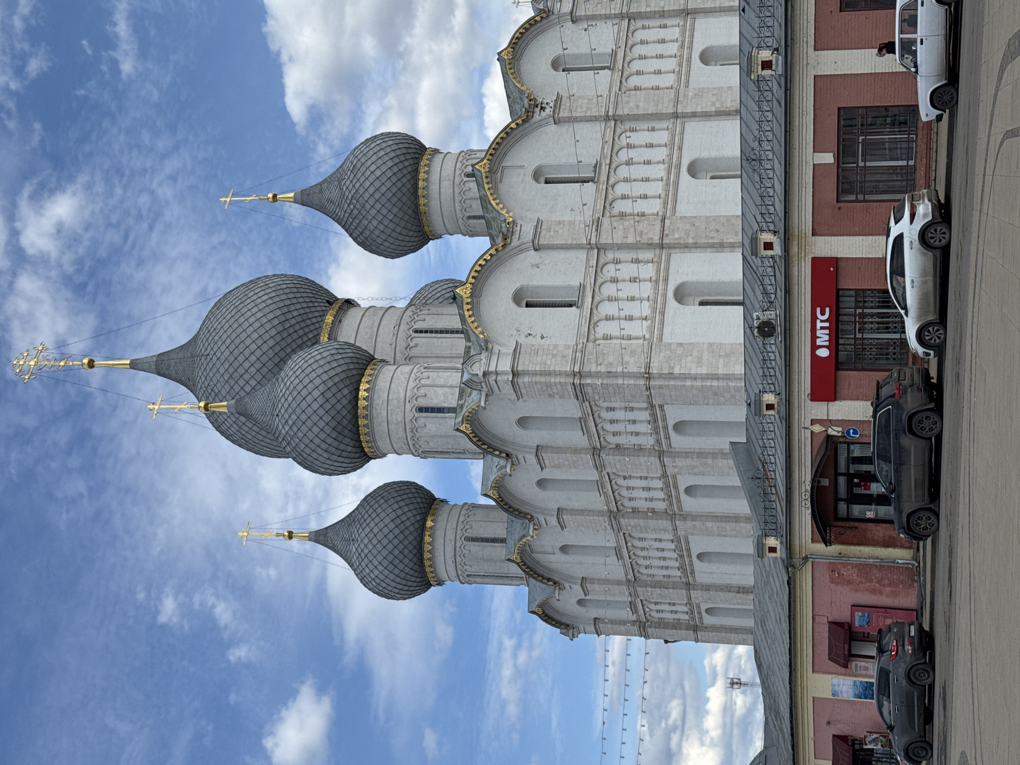 The image shows a building in Ново-Никольское, Russia, specifically the church of St. John the Baptist. The church has multiple onion-shaped domes, each topped with a cross. The facade of the church is decorated with arched windows and decorative elements. Adjacent to the church is a building with a red MTC sign. Several cars are parked on the street in front of the building. The sky above is blue with scattered clouds.