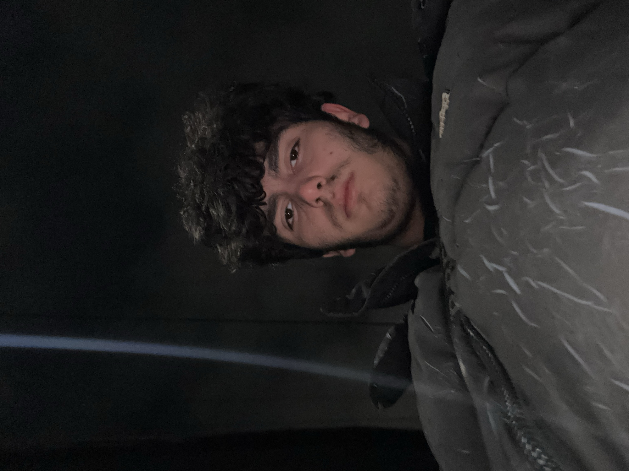 The image depicts a person with dark curly hair, brown eyes, and a beard stubble. The person appears to be wearing a dark-colored jacket. The background is predominantly dark, with a streak of light. The photograph seems to be taken at night. It is not possible to determine the location as Yerevan, Armenia from the image.