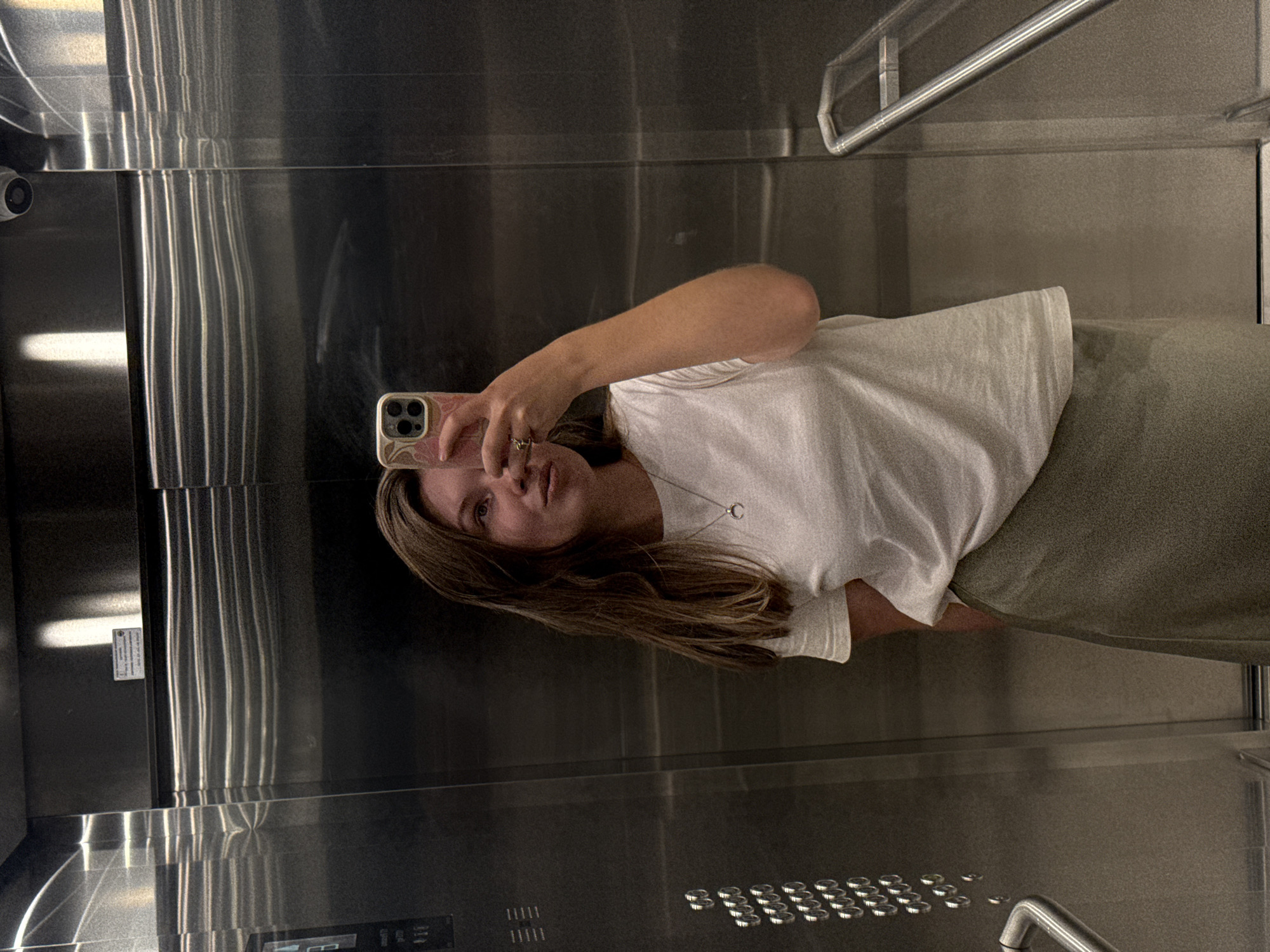 The image is a medium-shot, indoor, full-body selfie taken inside an elevator. A young woman with shoulder-length brown hair is standing in front of a brushed metal wall, holding a pink-cased smartphone in her right hand to take the photo. She is wearing a white t-shirt and a muted green skirt. She has a necklace with a crescent moon pendant. The elevator's interior is visible, including stainless steel walls, a control panel with buttons on the left, and a handrail on the right. The lighting appears to be artificial, reflecting off the metallic surfaces. The location is not specified in the image, and therefore cannot be determined to be Belgrade, Serbia.