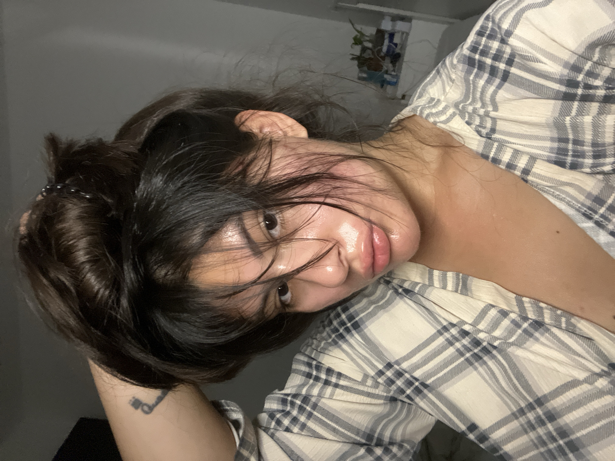 A young woman with dark hair pulled up in a messy bun is holding her hair with her left hand. She is wearing a plaid shirt. Her face is shiny and appears to be sweating or have a product on it. She has a tattoo on her left arm. The background is a plain wall, suggesting an indoor setting, possibly a bedroom or bathroom. There are no identifiable landmarks or features that would indicate a specific location in Kiryat Bialik, Israel.