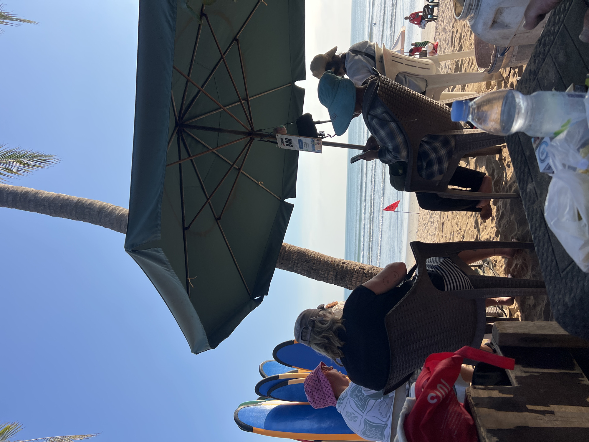 The image depicts a beach scene in Kuta, Indonesia, characterized by a clear blue sky and a tranquil ocean. In the foreground, several individuals are seated in chairs under a large green umbrella, suggesting a relaxed atmosphere at a beachside establishment. Some are engaged with their phones, while others appear to be conversing or simply enjoying the view. Surfboards are visible in the background, leaning against a palm tree, indicating the popularity of surfing in the area. A small red flag planted in the sand suggests a designated swimming or safety zone. The presence of a bar sign further confirms the location as a commercial establishment catering to beachgoers. The overall ambiance is one of leisure and enjoyment of a tropical beach environment.