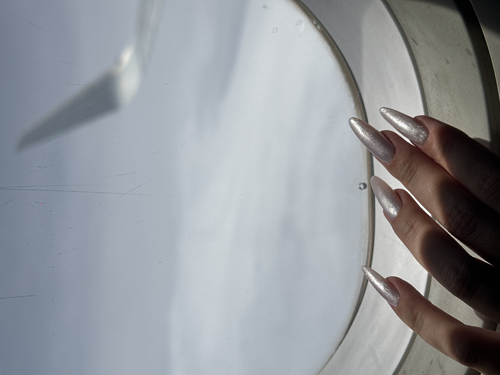 The image shows a close-up of a person's hand with long, pointed, light-colored, glittery nails resting on the edge of an airplane window. The window frame is white and rounded, and the view outside appears to be clouds, with a blurred airplane wing visible in the upper right portion of the frame. The lighting suggests it is daytime. The scene is consistent with being inside an airplane. No specific location within the city of Antung Vien, Cambodia can be determined from this image.