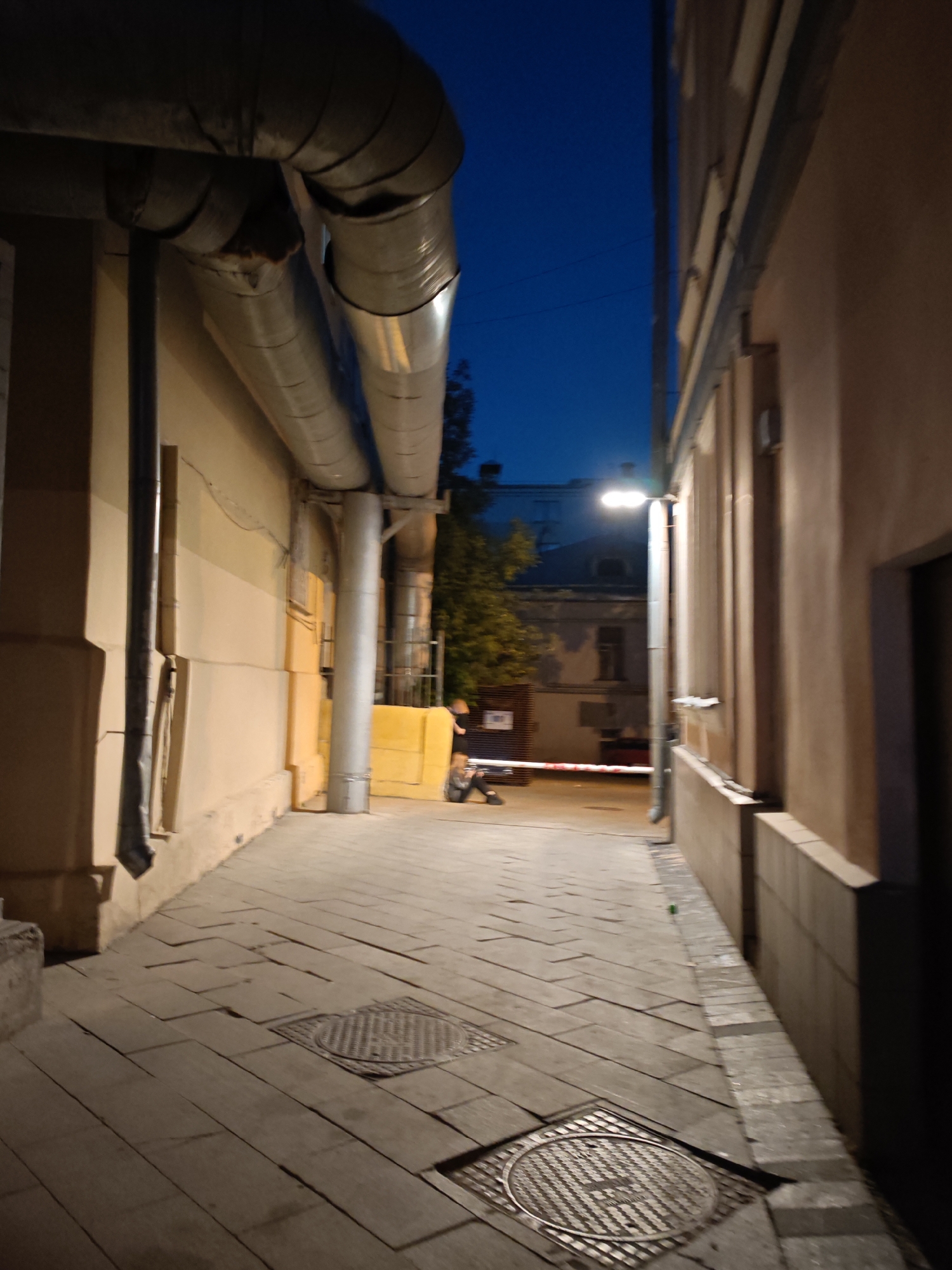 The image shows a narrow alley in Moscow, Russia, at dusk or night. The alley is paved with rectangular stones and has two circular manhole covers. Large, insulated pipes run along the left side of the alley, supported by vertical posts. The walls of the buildings on either side of the alley are beige/light yellow. At the end of the alley, a barrier is visible. A person is sitting on the left side of the alley near the barrier. There is a street light illuminating the scene. The sky is a dark blue color. The lighting is dim and mostly artificial.
