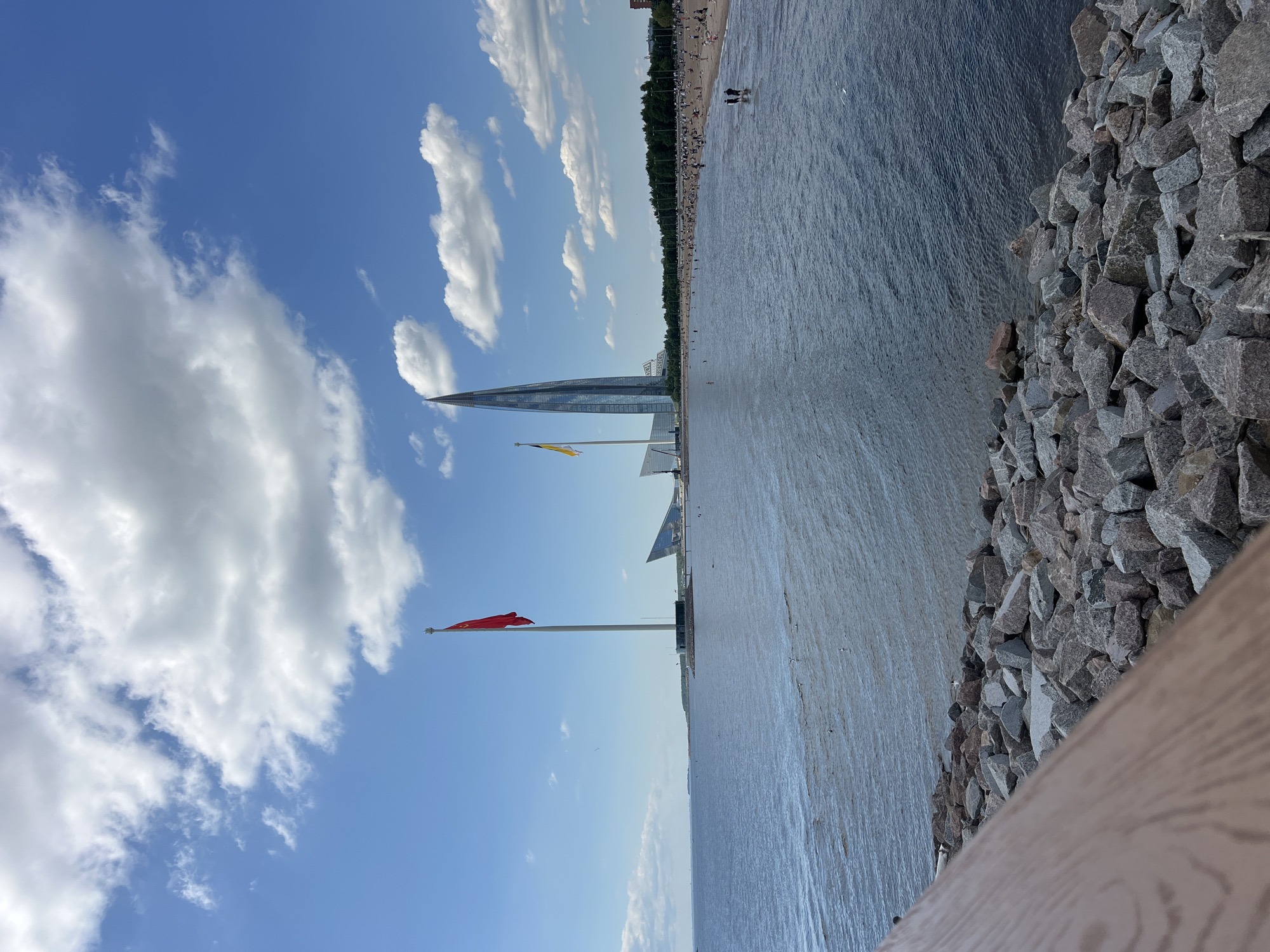 The image depicts a view of the Gulf of Finland from a rocky shoreline in St. Petersburg, Russia. In the foreground, a wooden railing runs diagonally from the bottom left corner. The middle ground features a calm body of water with ripples, and a shoreline dotted with people sunbathing and walking. Two prominent flags fly on tall poles in the water. The left flag is red, and the right flag is yellow and blue. In the background, a modern, glass skyscraper with a distinctive pointed top, the Lakhta Center, dominates the skyline, along with other contemporary buildings. The sky is a vibrant blue with scattered white cumulus clouds, suggesting a clear and sunny day.