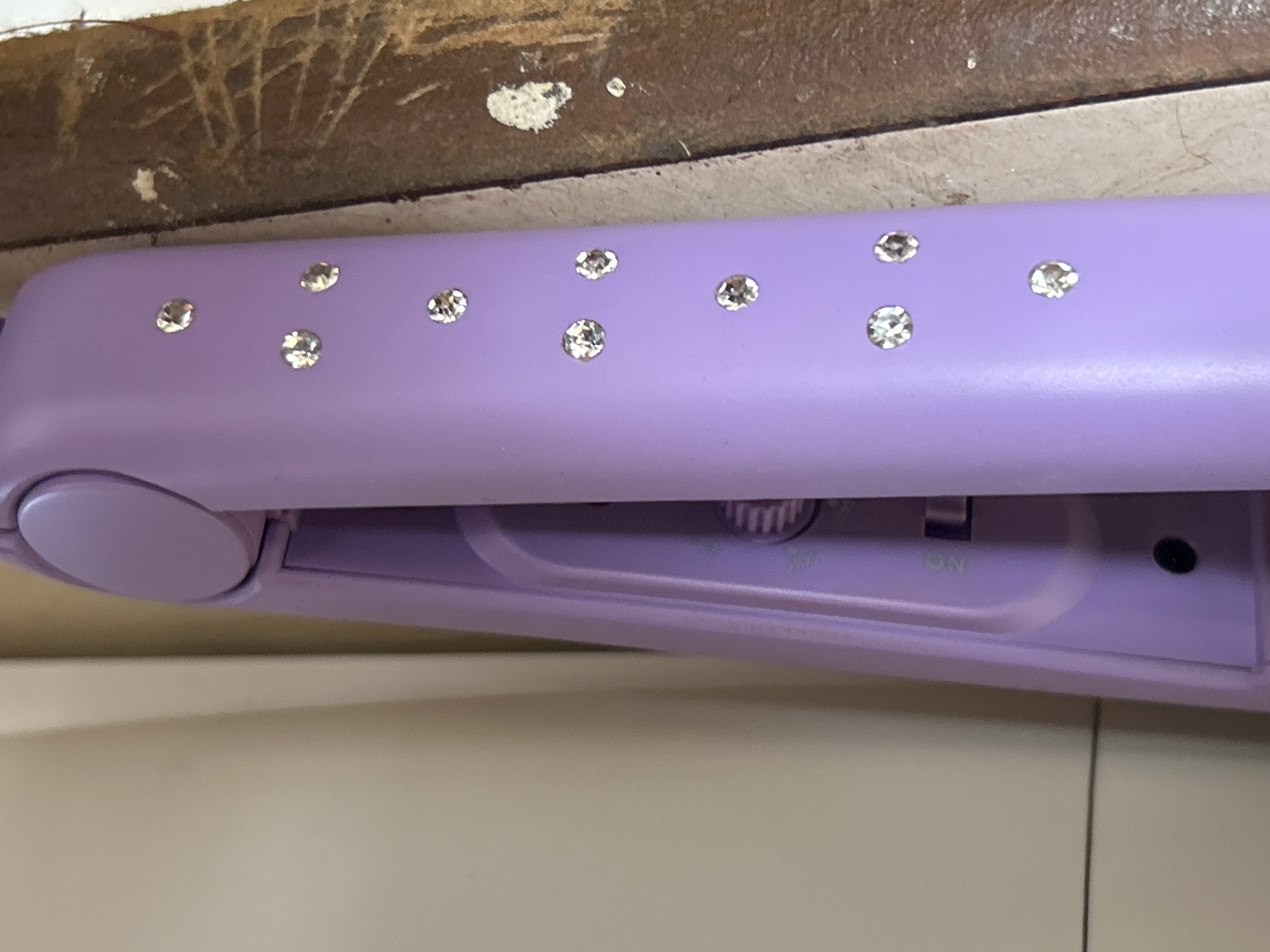 A close-up, eye-level shot shows a lavender hair straightener decorated with small rhinestones. The hair straightener is resting on a textured, brown surface. The background is a soft, light-colored surface, possibly a wall or shelf. The scene is indoors.