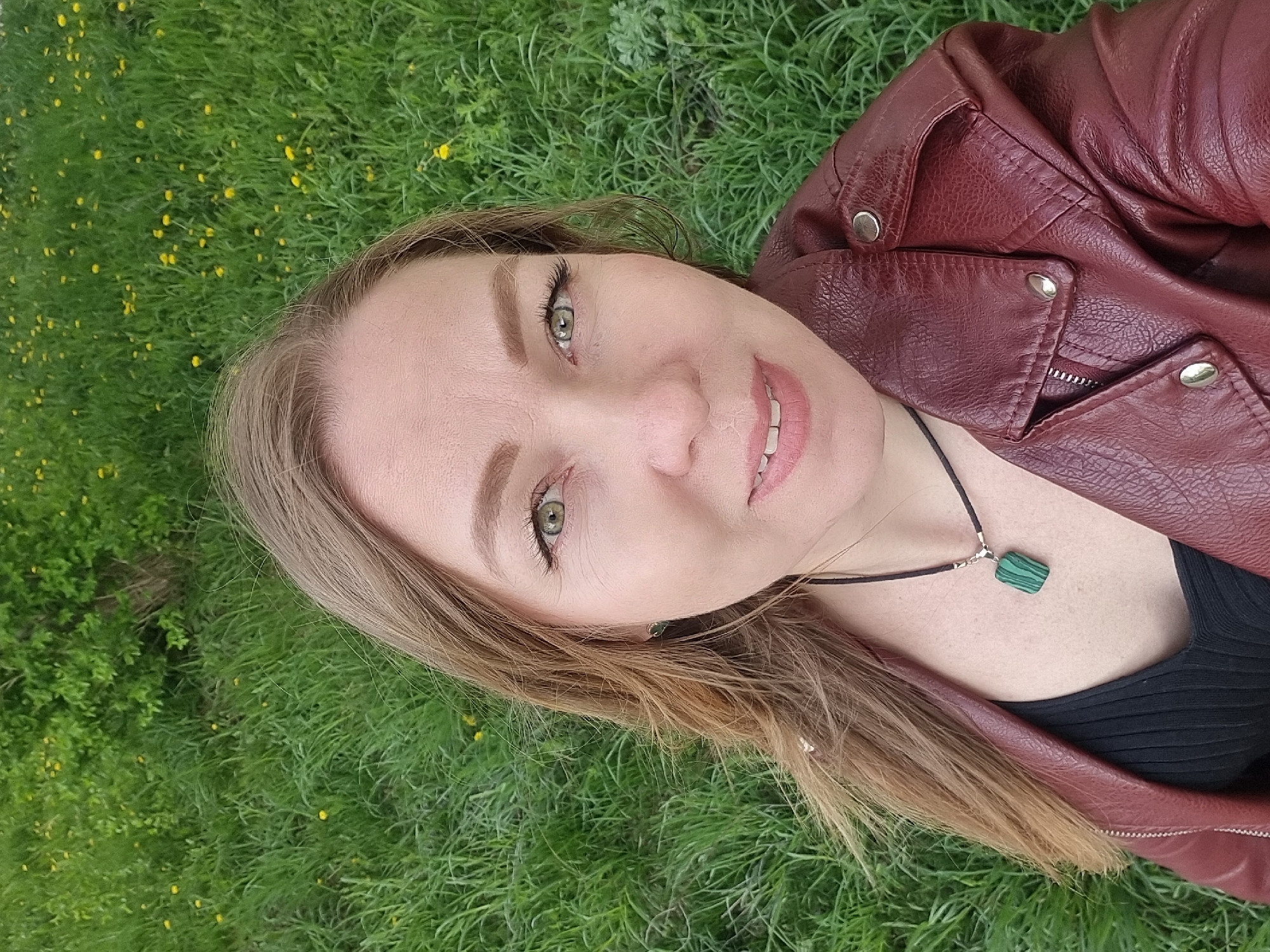 A woman with blonde hair and green eyes is lying on a grassy field with small yellow flowers. She is wearing a dark red leather jacket and a black top. A black necklace with a rectangular green pendant is visible on her chest. The background is a green field with small yellow flowers. This is in Chaikovsky, Russia.