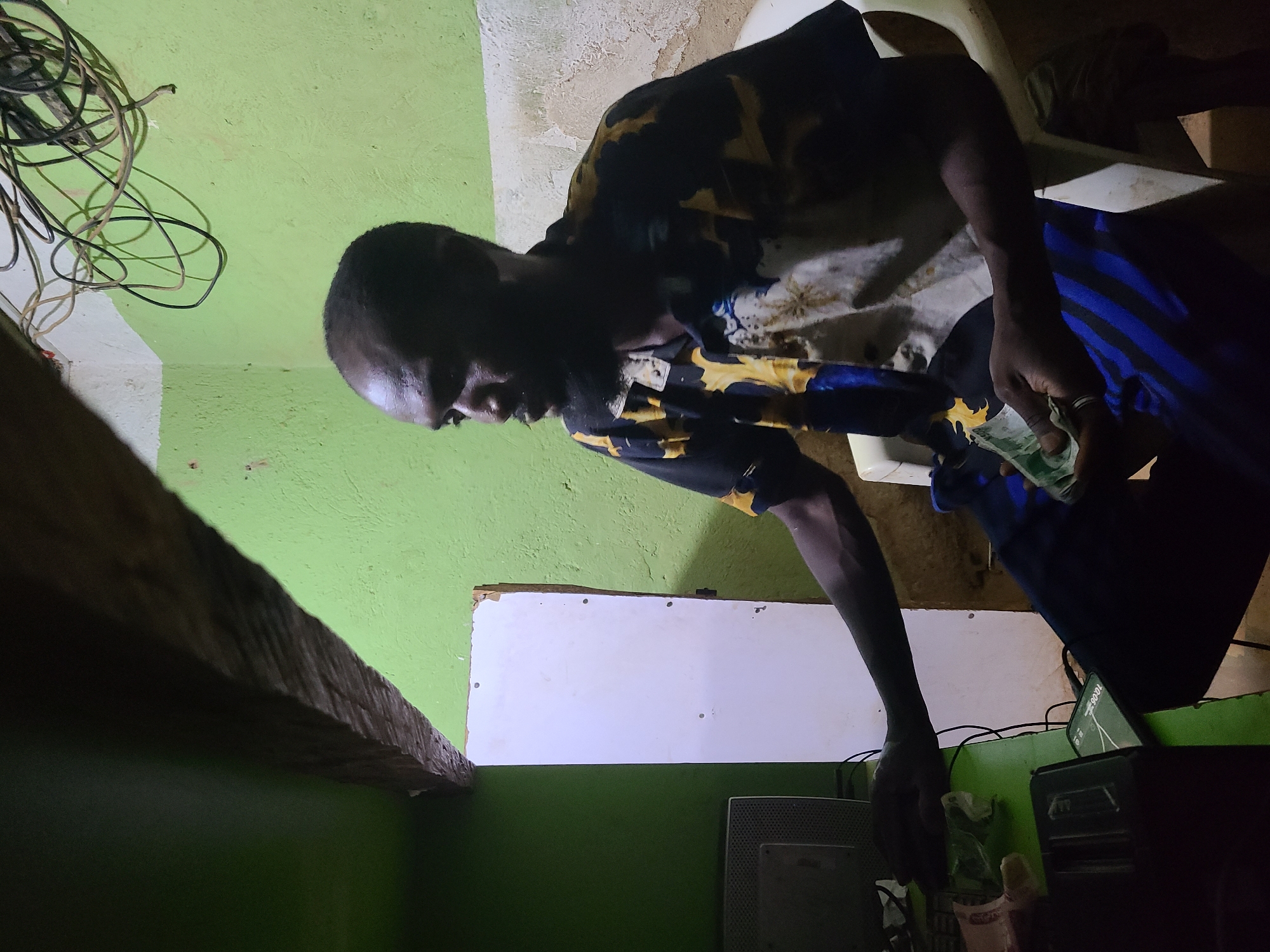 A man stands in a room in Jos, Nigeria. The walls are green with a white patch. The man is holding money in his right hand and wearing a patterned shirt and blue pants. There are wires hanging in the corner of the room. A wooden beam is partially visible in the lower left. A white object is visible on the wall. A computer monitor and other electronic devices are on a surface in the lower right.