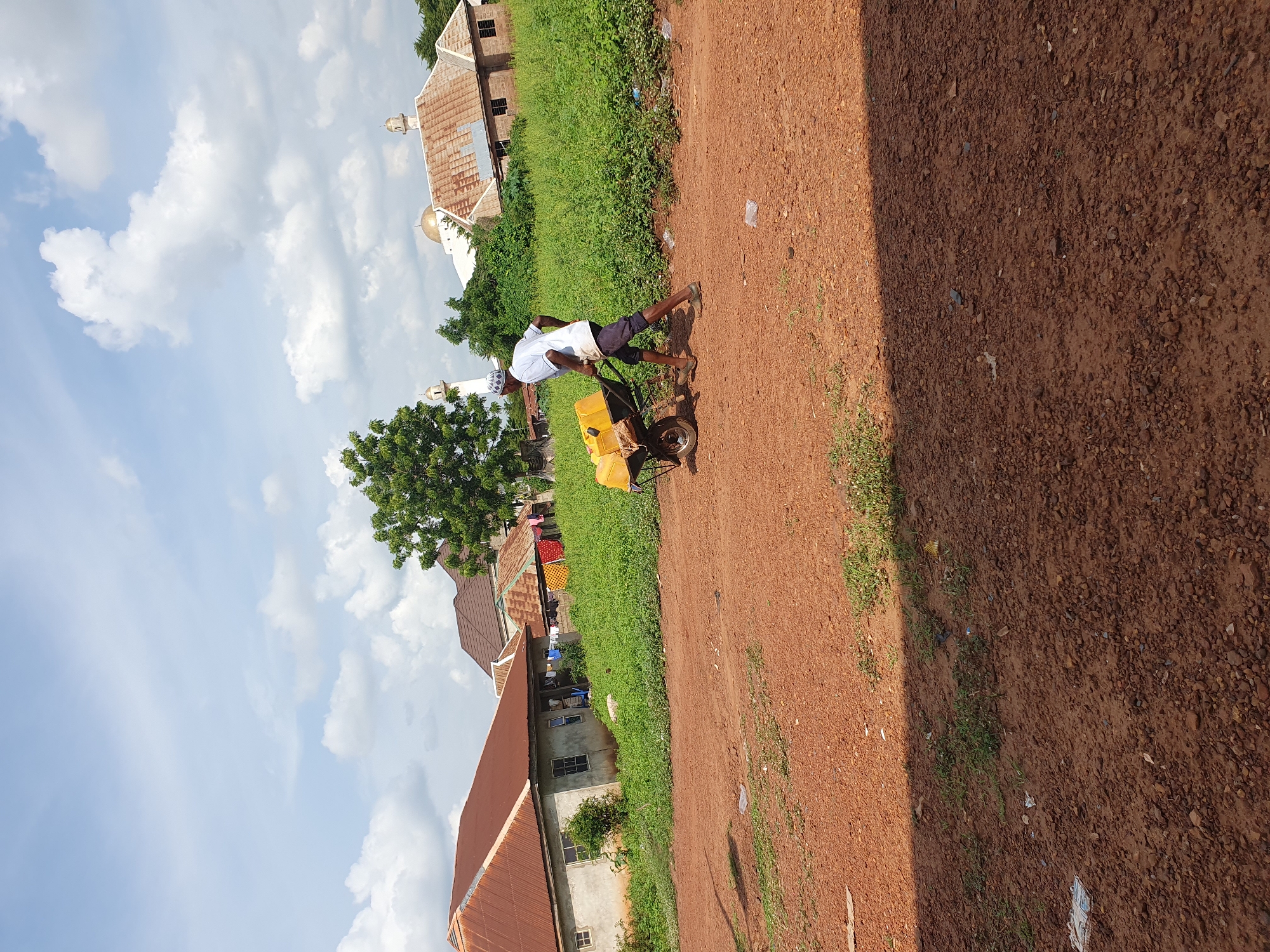 The image, originally rotated 90 degrees counter-clockwise, displays a daytime scene in what appears to be a rural or semi-urban area of Idah, Nigeria. The central subject is a man, dressed in a white short-sleeved shirt, dark shorts, and a white cap, actively pushing a yellow wheelbarrow. The wheelbarrow is loaded with several yellow jerrycans, suggesting he is transporting water or another liquid.
The man is situated on a reddish-brown dirt path or unpaved road, which occupies a significant portion of the foreground and midground. The path is uneven, covered with small stones and dirt, with patches of green grass and weeds growing along its sides. Sunlight casts shadows on the ground, indicating a bright, sunny day.
In the background, on either side of the path, various buildings are visible amidst green trees and vegetation. To the man's right, a structure with a golden dome and a white minaret, characteristic of a mosque, stands out. Another building with a white minaret is visible further to the left. Other houses with reddish-brown corrugated iron roofs and light-colored walls are scattered throughout the background. The sky above is clear blue with numerous white, fluffy clouds. There is no discernible text in the image.