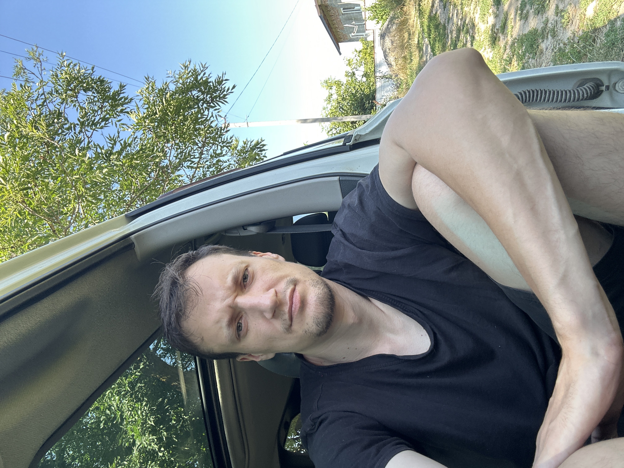 A man with short brown hair and a light beard is sitting in the open driver's side door of a white car, looking directly at the camera. He is wearing a black t-shirt and black shorts. His left arm is bent and resting on his left leg, and his right arm is bent at the elbow and resting on his leg. The car's sunroof is open, revealing green trees and a clear blue sky. In the background, there is a rural scene with a house, trees, a power pole, and power lines. The ground is covered with dry grass and dirt. The image appears to have been taken in Makiv, Ukraine, on a sunny day.