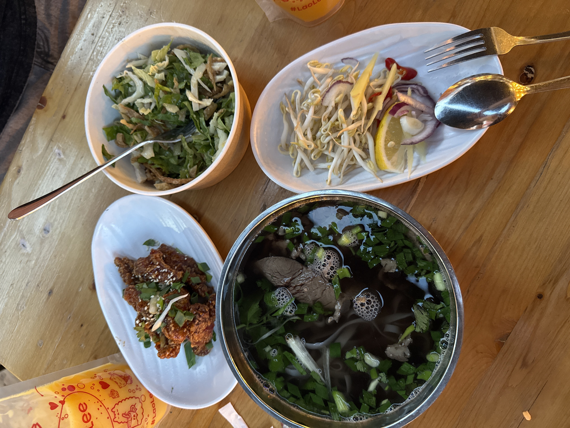 The image shows a wooden table with several dishes of food. There's a bowl with a green salad, a plate with bean sprouts, red onion, ginger, chili and a lemon wedge, a plate with fried chicken garnished with sesame seeds and greens, and a bowl of pho soup with beef and herbs. A fork and spoon are on the plate with bean sprouts. The setting suggests a casual dining environment. The location is not identifiable as Moscow, Russia, from the visual information.