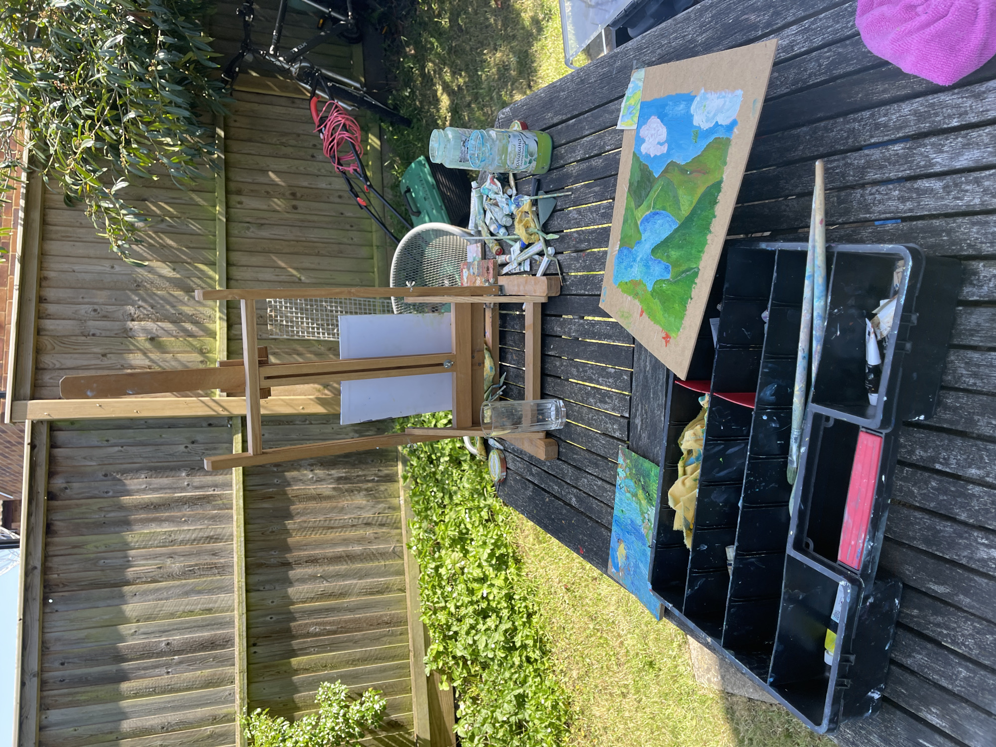 The image captures an outdoor painting setup in a garden in Maidstone, United Kingdom. An easel stands with two blank canvases, positioned near a weathered wooden picnic table laden with art supplies. Paint tubes and a glass jar with a green liquid sit amidst a partly finished landscape painting, depicting mountains, trees, a lake, and clouds. A black compartmentalized container, presumably for brushes and painting tools, occupies another section of the table. A lawnmower, fan, and other garden tools can be observed in the background. The environment includes a wooden fence, greenery, and what appears to be a section of a well-maintained lawn. The scene appears to be bathed in sunlight. There is no evidence of people or actions taking place in the scene.