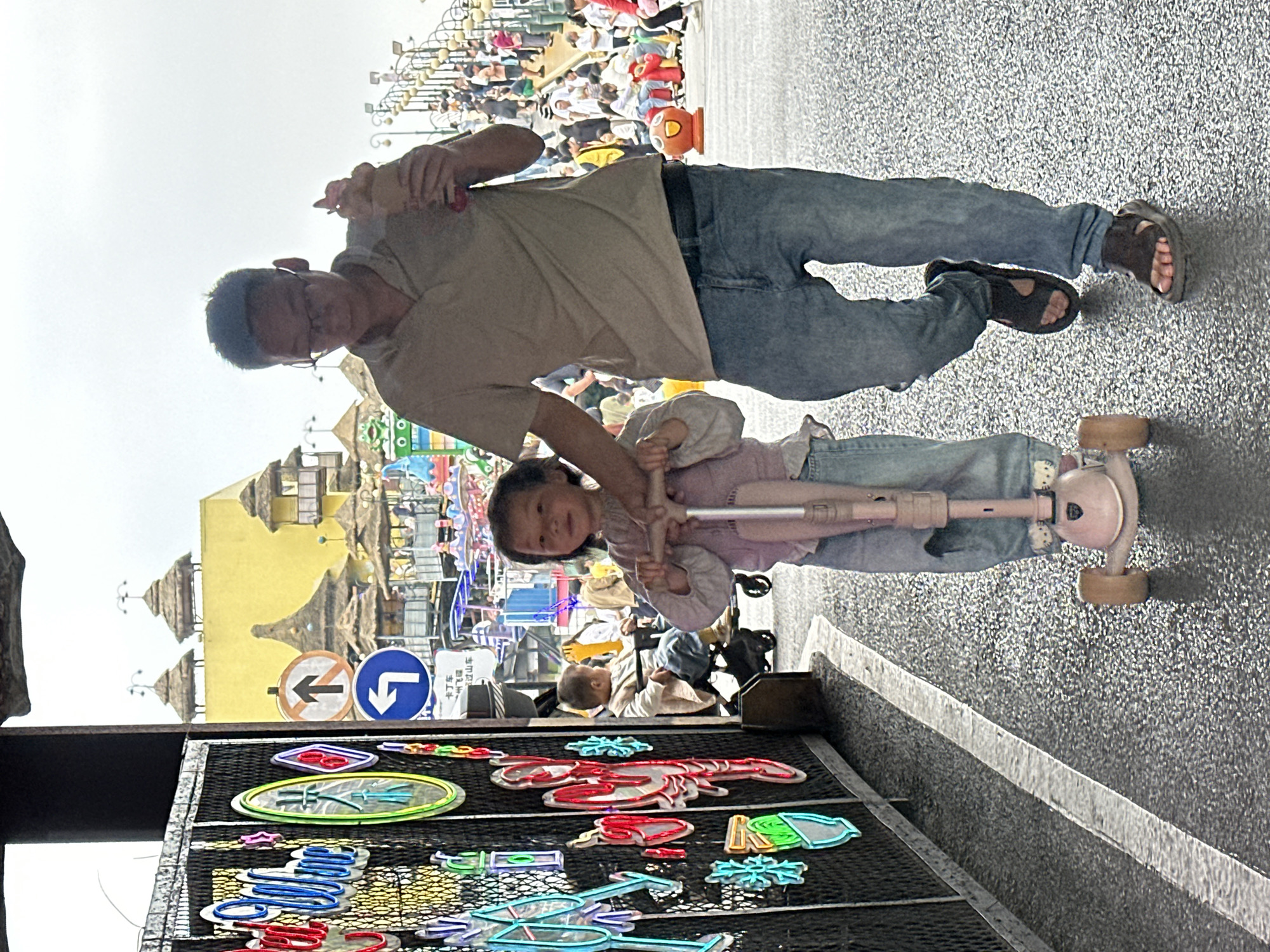 This vertically oriented image captures a man and a young girl engaged in a casual outdoor activity, likely within a public recreational area in Nan'an District, China. The man, wearing a light-colored short-sleeved shirt, blue jeans, and dark sandals, stands with a beverage in his left hand while his right arm supports a young girl. He wears glasses and has short dark hair. The girl, dressed in a light-colored top and jeans, is positioned on a pink and white three-wheeled scooter, gripping its handlebars. Both subjects are looking towards the camera with subtle smiles, suggesting contentment.
The background reveals a bustling scene with many people, indicative of a crowded public space like an amusement park or pedestrian street. Distinctive yellow buildings with ornate, possibly thatched, roofs are visible, adding character to the setting. In the lower left foreground, a display rack features colorful, neon-style cutouts depicting various shapes, including a prominent red lobster, blue snowflakes, and other abstract designs.
Mid-ground details include two circular road signs: one red-rimmed sign with a black arrow pointing left and a red diagonal slash, displaying the Chinese characters "禁止左转" (No Left Turn). Adjacent to it is a blue circular sign with a white arrow pointing downwards. The ground is paved with a speckled grey material. The bright, overcast sky indicates it is daytime. The overall atmosphere is lively and relaxed, capturing a moment of family leisure.