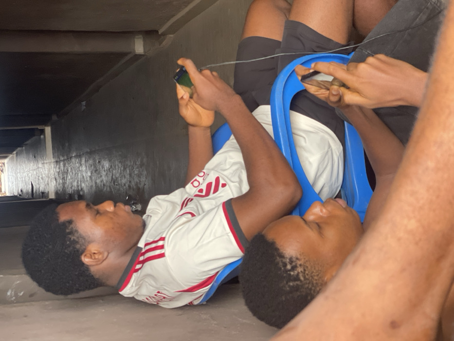 Two young black men lie on the ground under a dark structure, possibly a bridge or overpass. One wears a white and red soccer jersey, the other a white t-shirt and dark shorts. They sit in blue plastic chairs, appearing cramped and close together. Each holds a smartphone and seems to be engrossed in playing games. A charging cable dangles between them. The scene suggests a makeshift gaming spot in an unusual or less than ideal location.