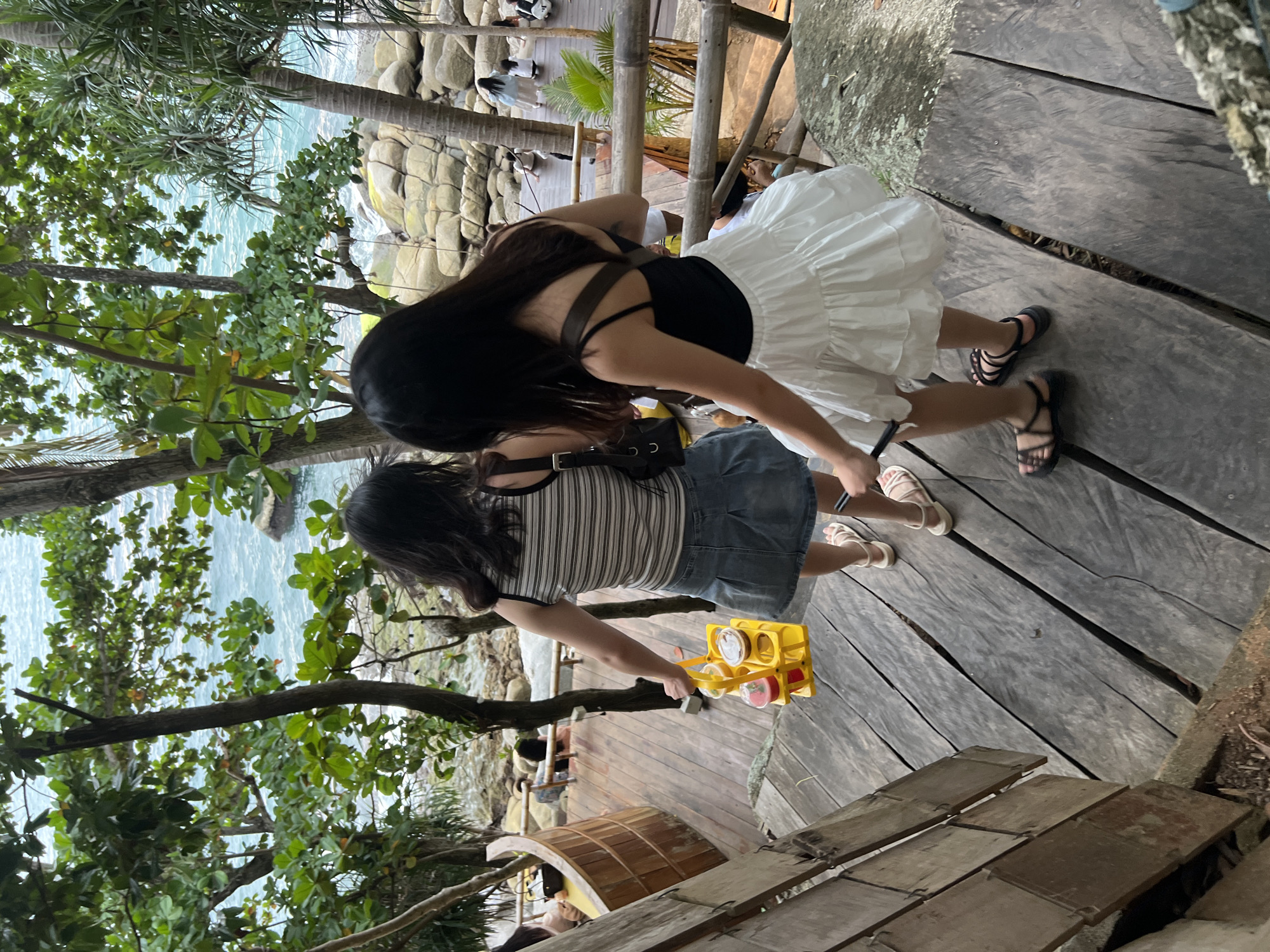 The image depicts two individuals on a wooden walkway near a rocky coastline. The person in the foreground is wearing a black and white striped t-shirt, a denim skirt, and sandals. They are holding a yellow carrier with drinks. The second person, slightly behind and to the right, is wearing a black top and a white tiered skirt, also with sandals. They are facing away from the camera.
The setting appears to be a tropical location, with lush green trees and the turquoise ocean visible in the background. Large rocks are scattered along the coastline. The walkway is made of weathered wooden planks, with some bamboo railings. The lighting suggests it is daytime, likely bright given the exposure. The scene seems relaxed and casual, possibly a tourist area or a resort in Karon, Thailand, given the location context.