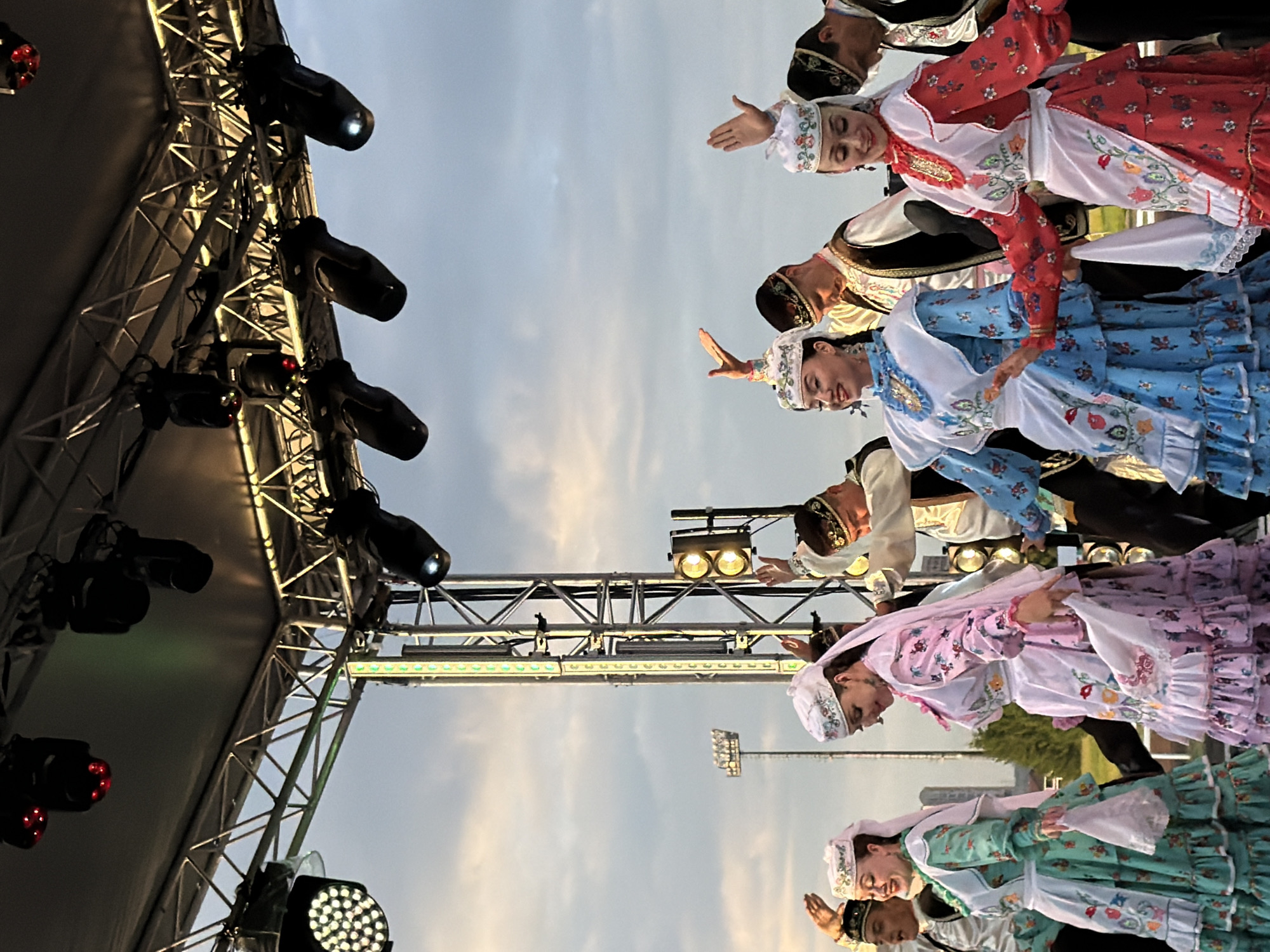 In the city of Unknown, Unknown, a group of dancers perform on an outdoor stage under a cloudy sky. The dancers, dressed in traditional colorful attire adorned with floral patterns, are engaged in a choreographed performance. Some wear intricately patterned hats, while others have veils. The stage is equipped with a lighting rig featuring several spotlights and LED strips, indicating an evening or dimly lit performance. In the background, tall buildings and stadium lights are visible, suggesting an urban or recreational setting. The dancers' hands are raised and their bodies are posed, suggesting movement and musical accompaniment.