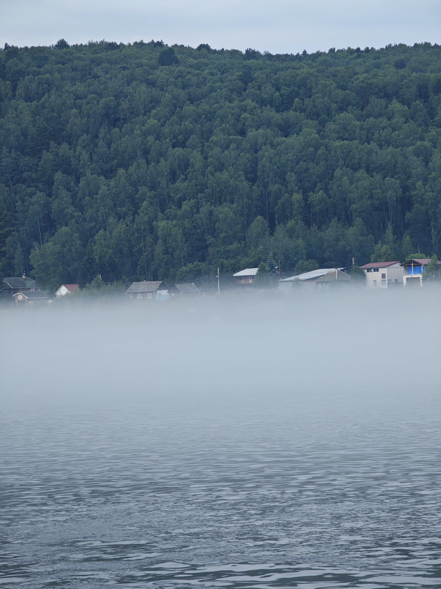 The image depicts a serene, misty landscape in the village of Atamanovo, Russia. In the foreground, a body of water, likely a river or lake, displays gentle ripples under a hazy sky. A dense, green forest blankets a hillside in the background. Nestled at the base of the hill, partially obscured by thick fog, is a small settlement of houses. The houses appear to be of various sizes and styles, with some featuring visible roofs and windows. The overall atmosphere is tranquil and somewhat mysterious due to the pervasive mist. No people or animals are visible in the scene.