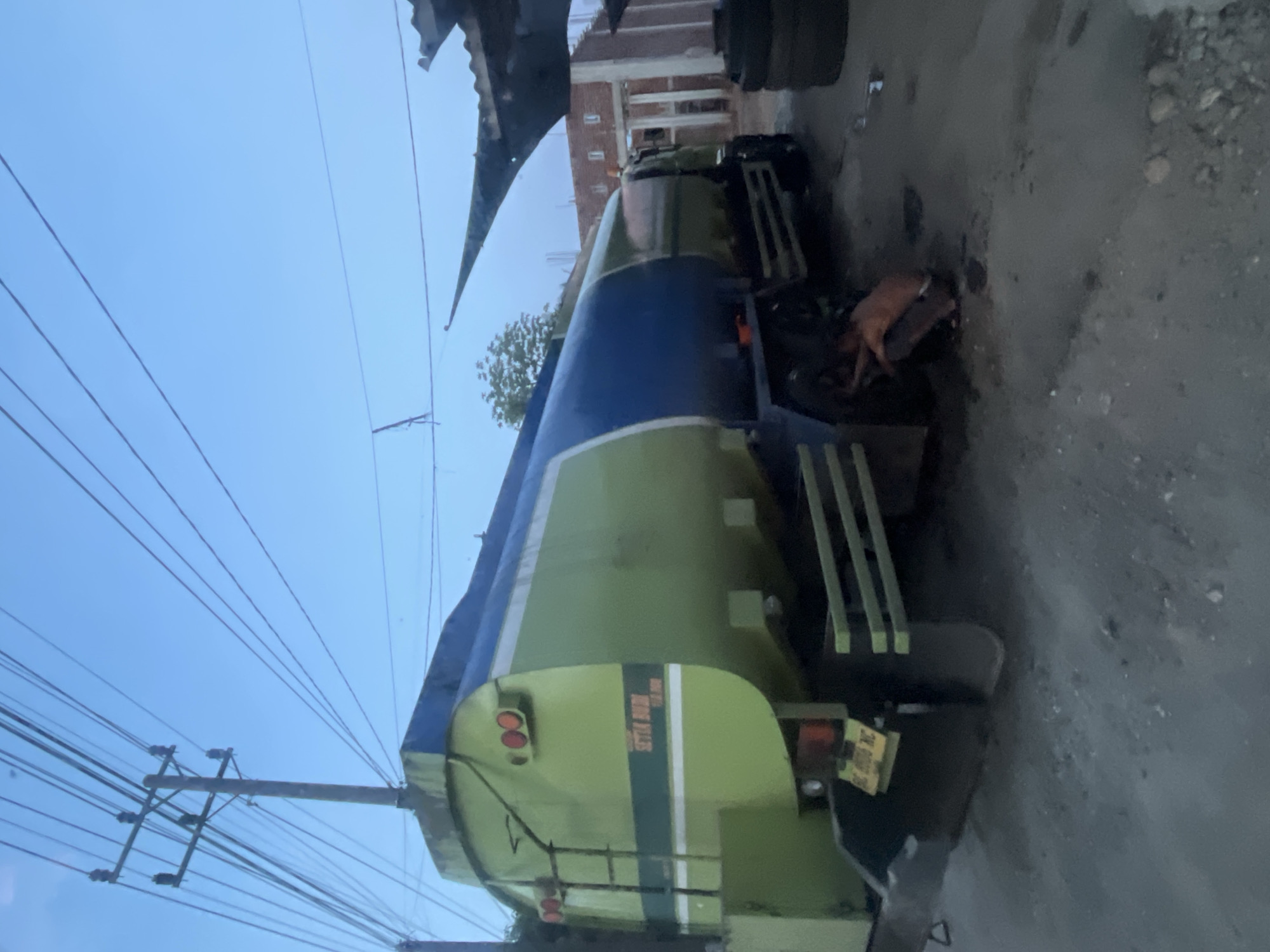 In Matangpanyang, Indonesia, an outdoor scene depicts a light green and blue tanker truck parked on a dirt or gravel lot. The truck is the primary subject, occupying the center and right side of the frame. A person, wearing an orange garment, is crouched beside the truck's rear passenger-side wheels. The truck has a ladder on its rear, and a license plate is visible. In the background, a partially constructed building with columns and a roof is present. Overhead, power lines stretch across the sky, with a utility pole to the left. The sky is a clear, pale blue, suggesting either early morning or late evening. A stack of tires is visible to the far right of the image.