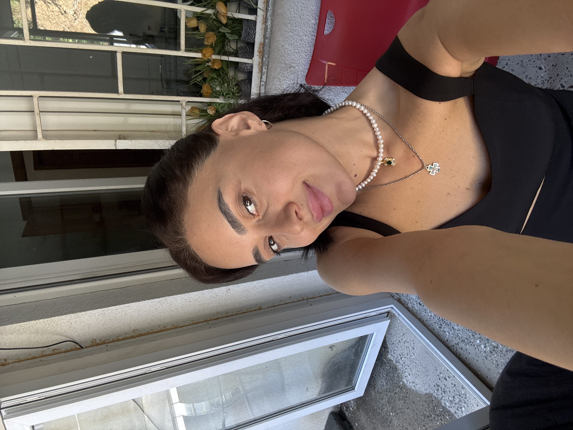 A selfie is taken by a woman with dark hair and fair skin. She is wearing a black top, a pearl necklace with a green pendant, and a silver cross necklace. She is smiling and looking at the camera. The background is blurred, but it appears to be an outdoor setting with a window and some plants. The woman is in Tbilisi, Georgia.