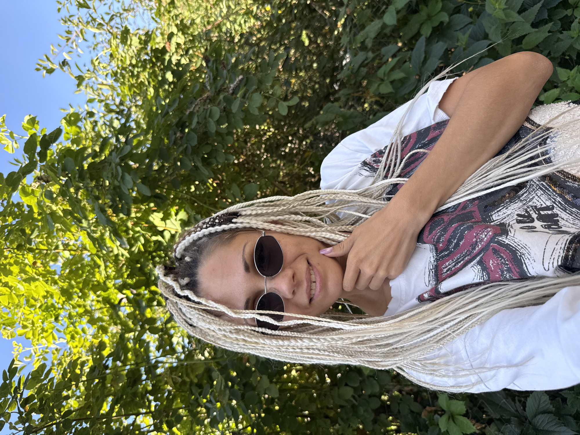 A medium shot captures a woman with blonde box braids and sunglasses, wearing a white t-shirt with a graphic print, standing outdoors. She is leaning her chin on her hand, smiling, and looking at the camera. Behind her, lush green foliage and a clear blue sky create a natural backdrop. The scene appears to be in the city of Rusanovo, Russia, possibly in a park or backyard area.