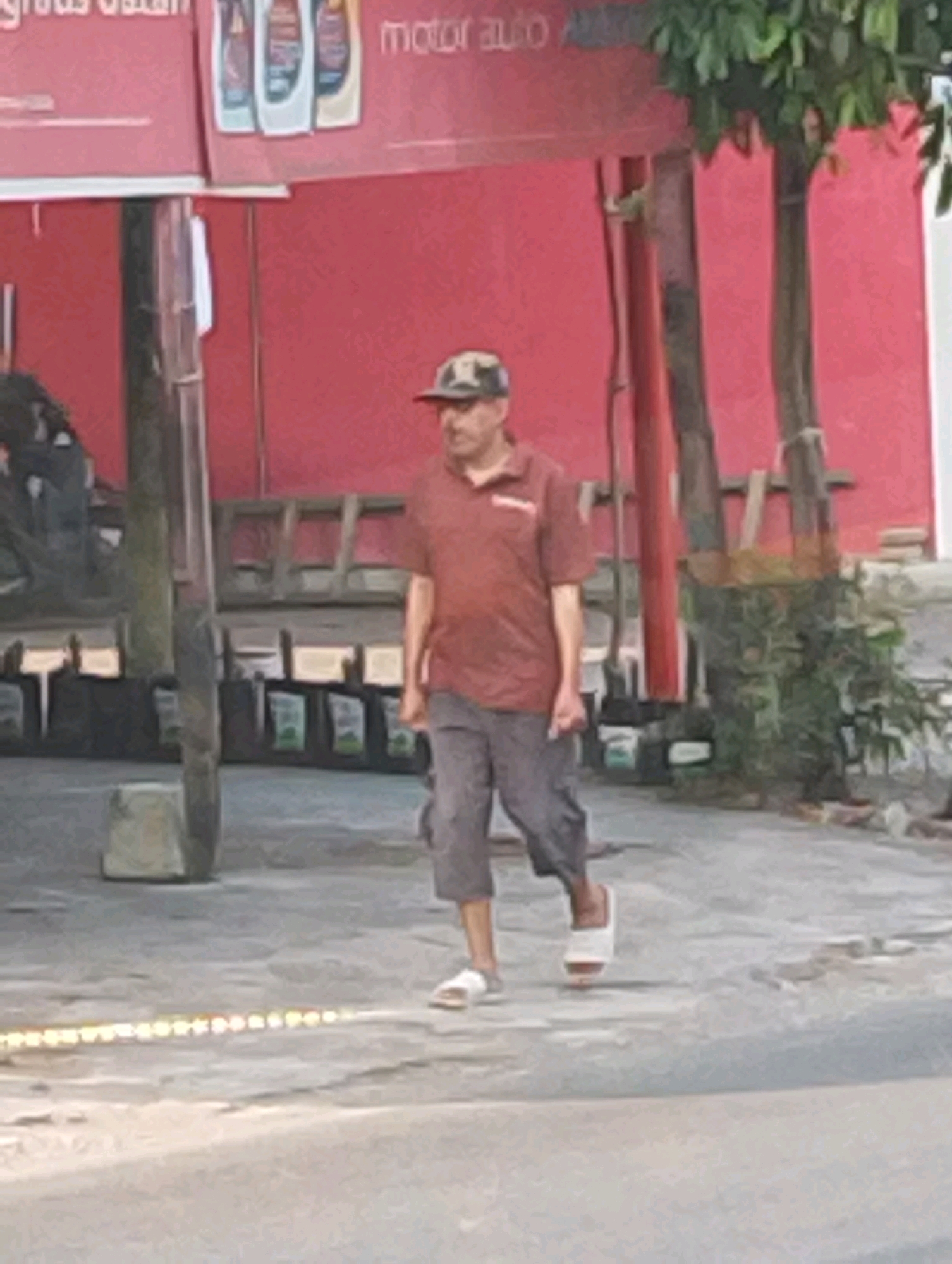 A man wearing a red polo shirt, grey pants, and a camouflage baseball cap walks on a paved surface in front of a red building. The building has signs that read "motor auto". There are several trees and some greenery visible. In the foreground, a line of lights or illuminated objects runs along the ground. The scene appears to be outdoors, possibly in a commercial area of Manaruwi, Indonesia.