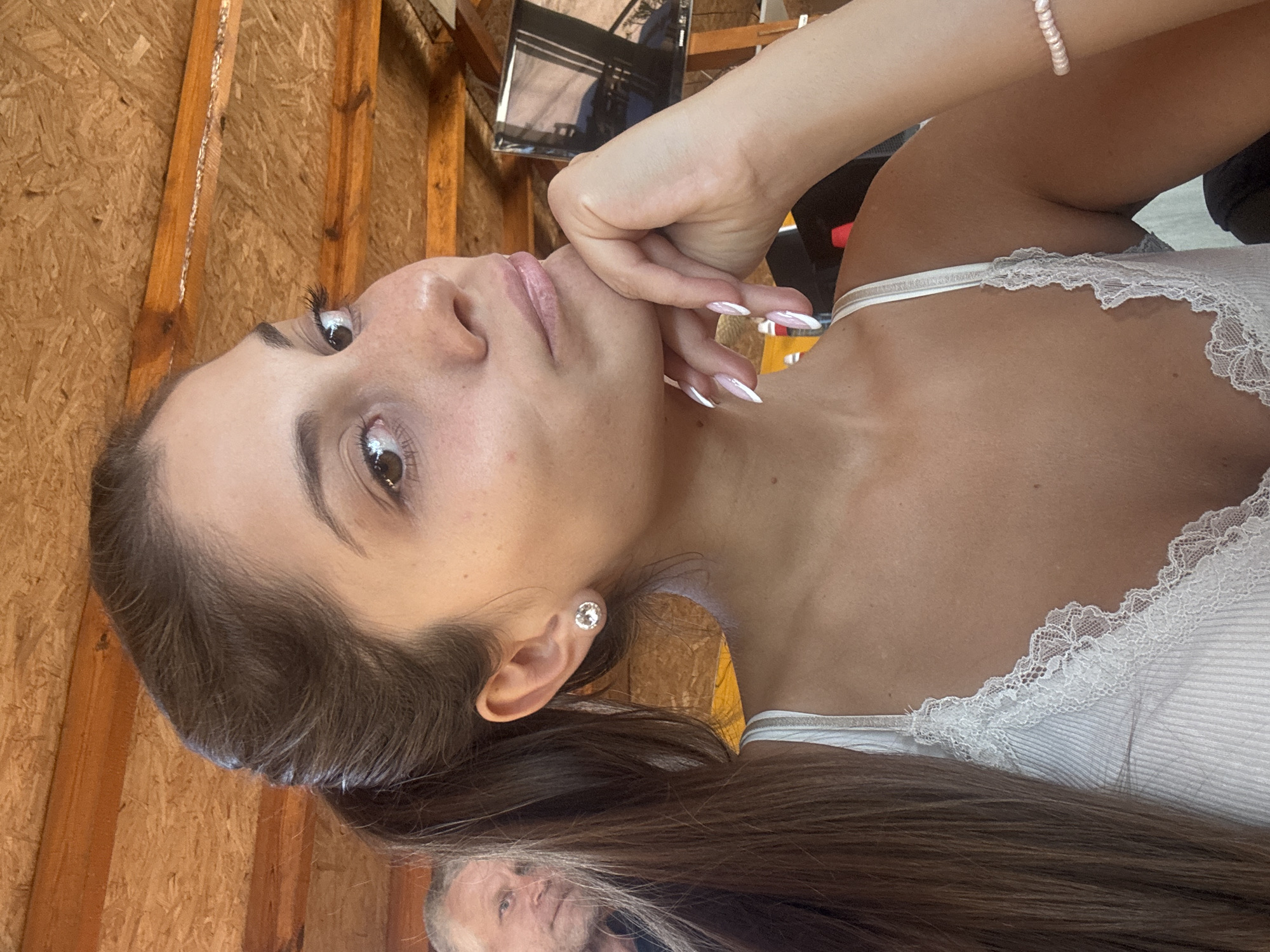 The image shows a woman with long brown hair, fair skin, and brown eyes. She is wearing a white tank top with lace trim. She has a bracelet on her wrist and an earring on her ear. Her hand is near her face. In the background, there is a wooden wall and a television. At the bottom of the image, there is a man's face. The location is not identifiable as Sozopol, Bulgaria from the provided image.