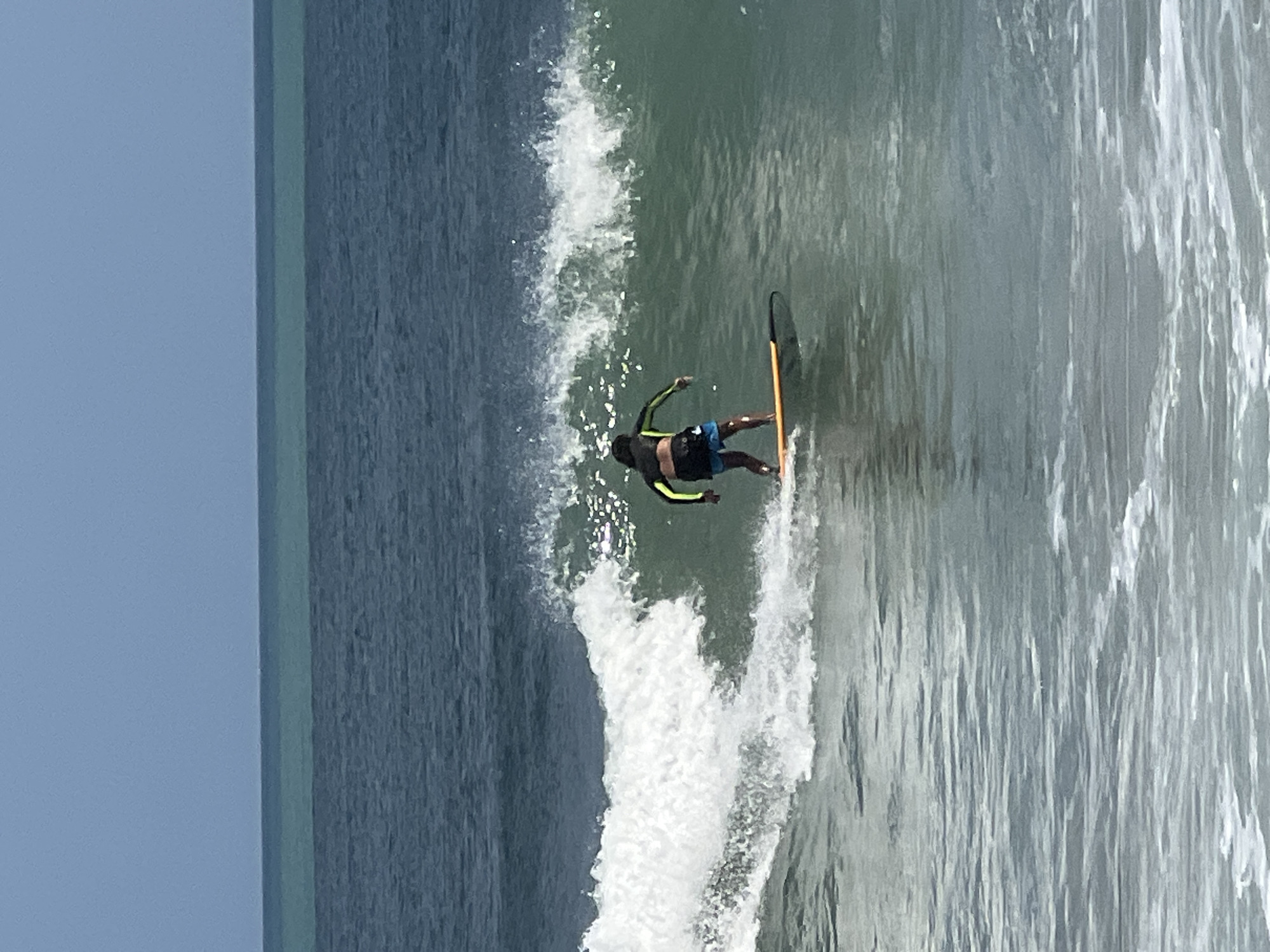 A person is surfing on a wave in the ocean. The surfer is wearing a black and yellow rashguard and blue board shorts and is standing on an orange surfboard. The wave is breaking to the left. The sky is clear and blue, and the ocean is a deep blue color. The scene is likely Kuta, Bali, Indonesia, a popular surfing destination.