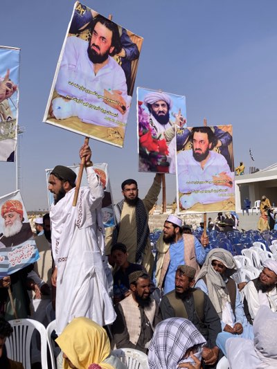 A public gathering is taking place outdoors under a clear blue sky in Pishin, Pakistan. A large crowd of men, predominantly adults, are present, many wearing traditional South Asian attire such as shalwar kameez, turbans, and prayer caps. Some men are seated on rows of white plastic chairs, while others stand, holding up large portraits.
The most prominent portraits feature a man with a dark beard and long dark hair, wearing a white shirt. Visible text on these banners identifies him as "مولانا ہدایت الرحمن بلوچ" (Maulana Hidayat-ur-Rehman Baloch). Below his image, additional text reads "ڈاکٹر شمعون مسیح کے خون سے روبرو ہو گی، اے ظالمان" (The blood of Dr. Shamoun Masih will confront you, O oppressors), suggesting a theme of protest or demand for justice. Another frequently displayed portrait shows a man with a white turban and white beard, often identified as Mullah Omar, with flowers in the foreground. A third portrait on the far left depicts an older man with a white beard and a red/orange turban, likely Maulana Fazal-ur-Rehman.
The bright sunlight indicates it is daytime. The arrangement of chairs and the display of political or religious figures suggest a rally, convention, or public address. The participants appear engaged and attentive.