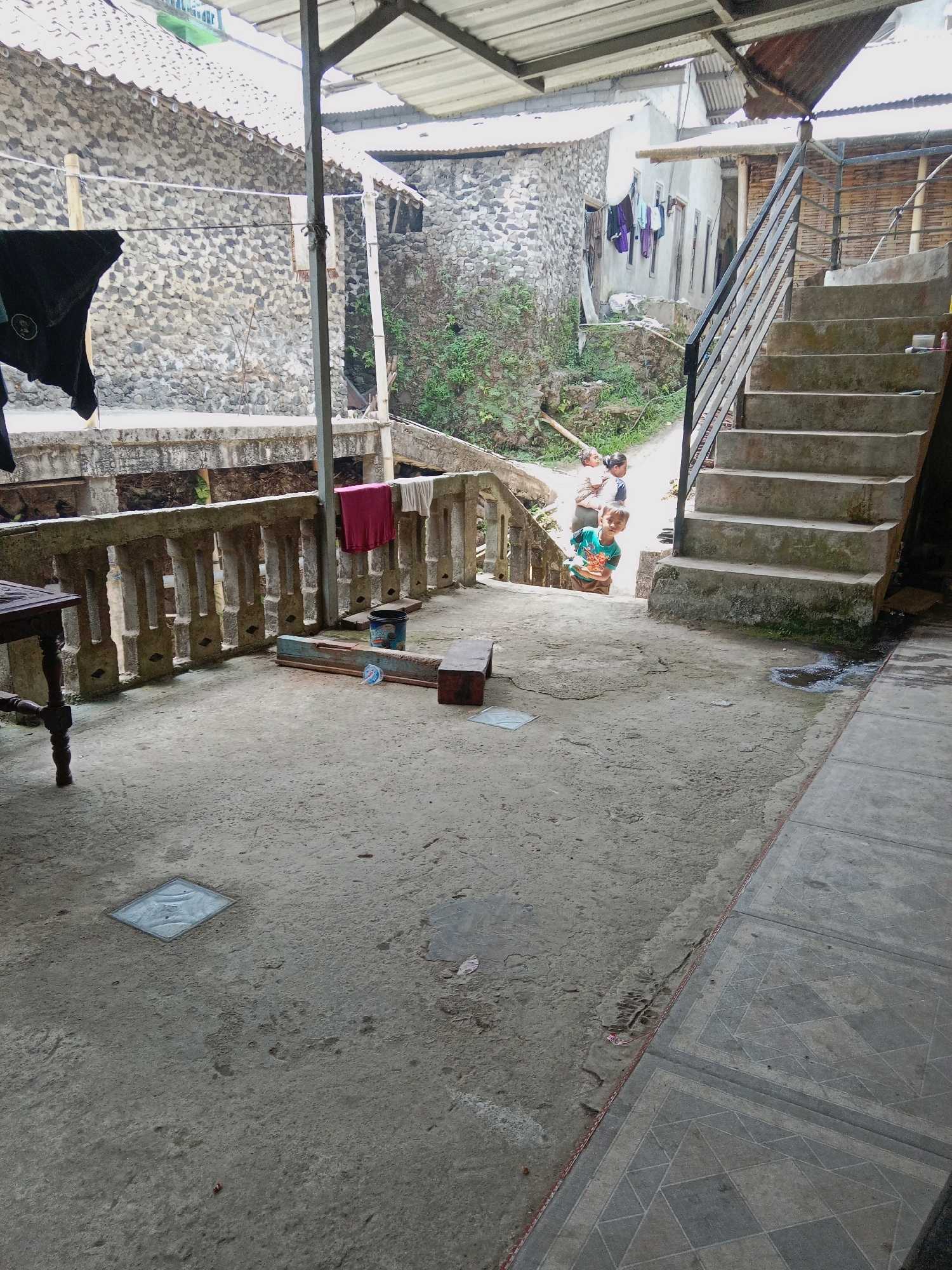 The image depicts an outdoor scene in Pulosaren, Indonesia, likely a residential area characterized by rough stone walls, concrete pathways, and a partially covered outdoor space. In the foreground, a concrete ground with a square metal grate is visible, and to the left, a dark wooden table with carved legs is positioned. A concrete railing with vertical supports runs along a raised area. Hanging on the railing are two pieces of cloth, one red and one white. Further back, a set of concrete stairs with a black metal handrail ascends to the right of the frame.
In the middle ground, three children are present. Two appear to be standing or walking, and one, a young boy in a green t-shirt, is looking directly at the camera. They are positioned on a path or lower level relative to the foreground.
The environment suggests a functional living space with elements of construction and everyday life. Laundry is seen hanging on a clothesline in the upper left, and a bucket and some wooden planks are on the ground near the railing. The architecture includes stone walls and metal and tin roofing. The overall impression is one of a somewhat rustic, utilitarian setting.
