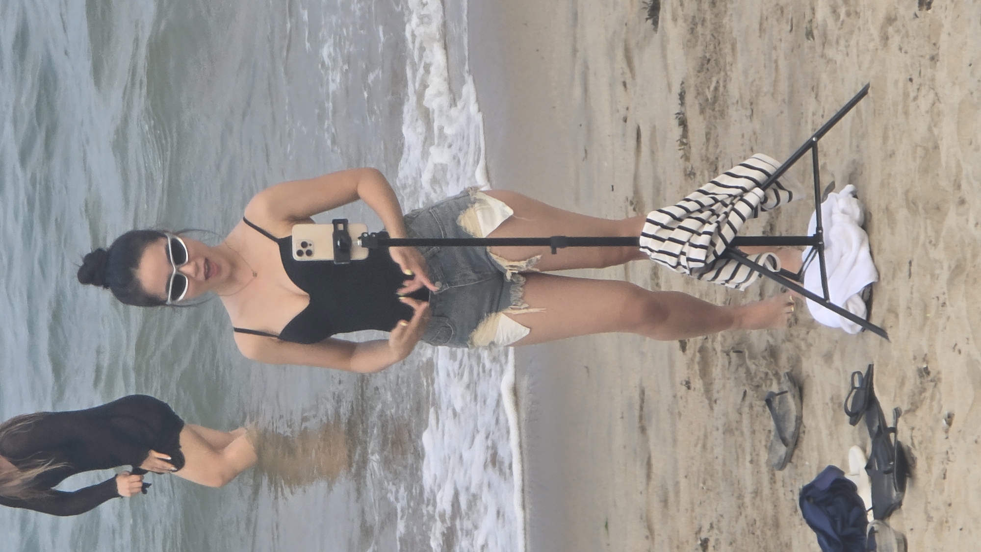 A young woman stands on a sandy beach at the edge of the ocean, posing in front of a smartphone mounted on a black tripod. She wears a black top or swimsuit, distressed denim shorts with white fabric showing through, and white-framed sunglasses. Her dark hair is pulled back in a bun. She looks towards the phone with her mouth slightly open, and her hands are positioned in a gesture as if for a photo or video. The phone, a light-colored iPhone, is held by a black clamp on the tripod.
Behind and slightly to her left, another person is partially submerged in the shallow ocean water, seen from the back and wearing a dark, long-sleeved garment. Gentle waves with white foam break on the shore behind the subjects.
On the sand next to the tripod, a black and white striped garment is draped over one leg, and a white towel or cloth rests on another. Nearby on the sand are two pairs of dark sandals or flip-flops and a folded dark blue fabric item.
The scene takes place during the daytime, under bright natural light, suggesting clear weather. The setting is a beach, specifically Nha Trang, Vietnam, with light-colored sand and clear water. The primary activity appears to be a personal photoshoot or video recording session. No discernible text is visible in the image.