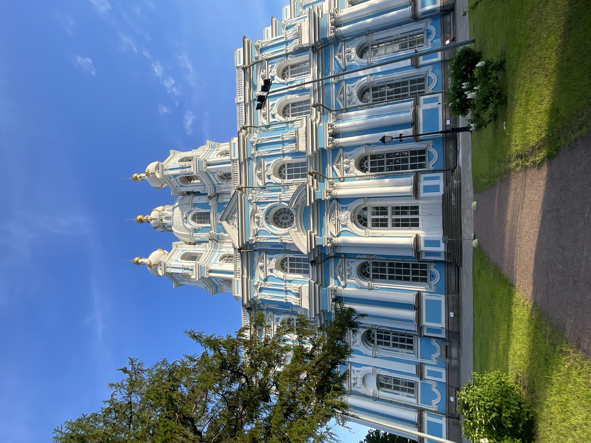 The image displays the Smolny Cathedral located in St. Petersburg, Russia. The cathedral, a prominent example of Russian Baroque architecture, features a multi-tiered facade rendered in sky-blue and white. White fluted columns, pilasters, and intricate stucco ornamentation frame numerous arched windows. Three white domes, each topped with a golden spire, are visible against the clear blue sky, which contains a few wispy white clouds.
In the foreground, a brown dirt path curves gently from the lower left to the lower right. It is bordered by a strip of vibrant green grass, which shows areas of both direct sunlight and shadow. A large evergreen tree with dense, dark green foliage occupies the left side of the frame, partially obscuring the lower left portion of the cathedral. Smaller green bushes are scattered on the grass, with one cluster on the right exhibiting white flowers. A black, ornate lamppost stands vertically on the right side of the path. Higher on the right, a tall black pole supports a multi-directional floodlight array, angled towards the cathedral. Small informational signs are visible on the ground near the base of the building. A light-colored, spherical object rests on the ground adjacent to the path on the far right. The scene is illuminated by strong daylight.