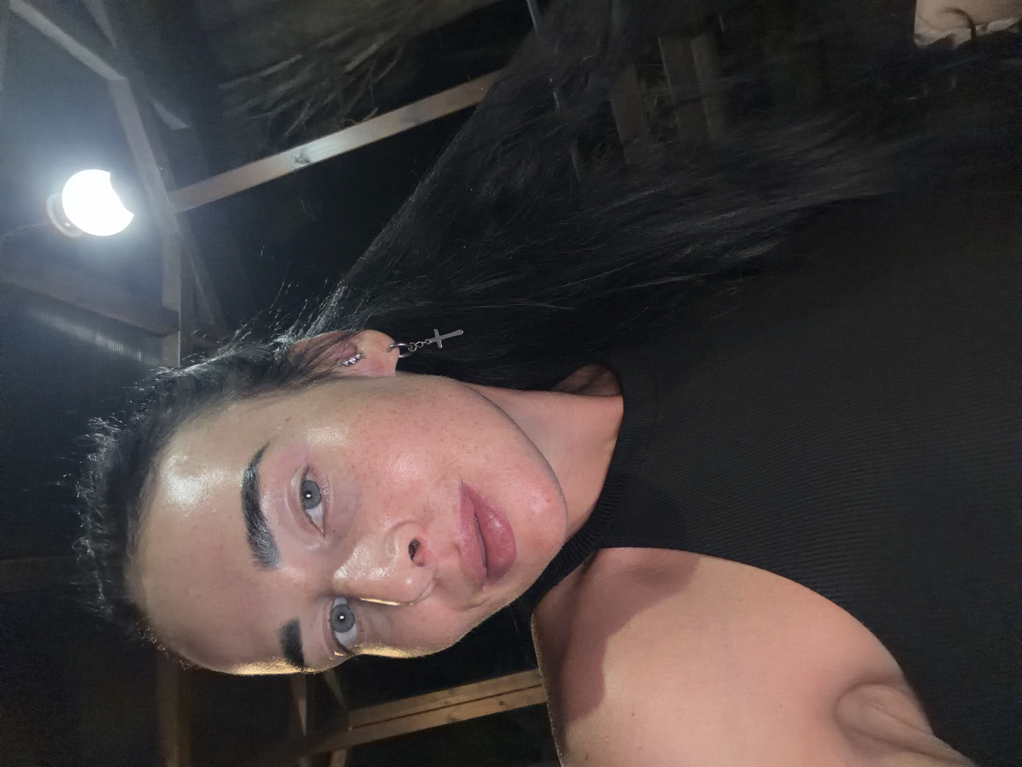 A woman with long, dark hair and light blue eyes is shown from the chest up in this close-up selfie. She is wearing a black sleeveless top and a silver earring with a cross pendant. Her skin appears to have some light freckling. The background is dark and out of focus, with a bright light fixture visible above and to the right of her head. The setting appears to be outdoors, possibly a patio or covered area, with wooden beams and supports visible in the background. There is no discernible information about the location being Sochi, Russia.