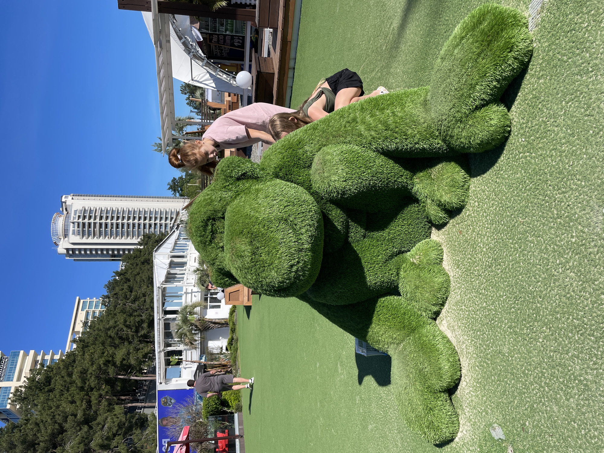 The image displays a lively outdoor scene on a bright, sunny day, likely in a resort or recreational area in Sochi, Russia. The ground is uniformly covered in artificial green turf, creating a manicured, park-like environment.
In the foreground, a prominent feature is a large topiary sculpture, shaped like an animal (possibly a dog or bear) in a lying posture. This sculpture is also made of artificial green grass, mirroring the ground cover. Two young individuals are actively engaging with the topiary. One, with reddish-brown hair tied up, wearing a pink t-shirt, leans over the sculpture's head. The second individual, with blonde hair, dressed in a dark green tank top and black shorts, is lying prone on the topiary's back, facing away, suggesting playful interaction.
Further back, an adult male in a dark grey t-shirt and shorts walks across the turf. The background is dominated by a clear blue sky and several buildings. A tall, modern white high-rise building with many windows stands prominently to the left. To the right, smaller white buildings, possibly cafes or pavilions, feature white awnings and wooden decking, indicative of outdoor dining or leisure spots. Tall evergreen trees are visible, interspersed among the structures, adding to the lush setting.
The scene is illuminated by bright midday sun, casting distinct shadows. The atmosphere appears relaxed and joyful. Visible text includes a partial word, possibly "BOULEVARD," on a building in the distance. In the lower-left background, a colorful mural with faces and some indistinct Cyrillic text can be seen, alongside a red object. The overall impression is one of leisure and enjoyment in a modern, coastal urban landscape.