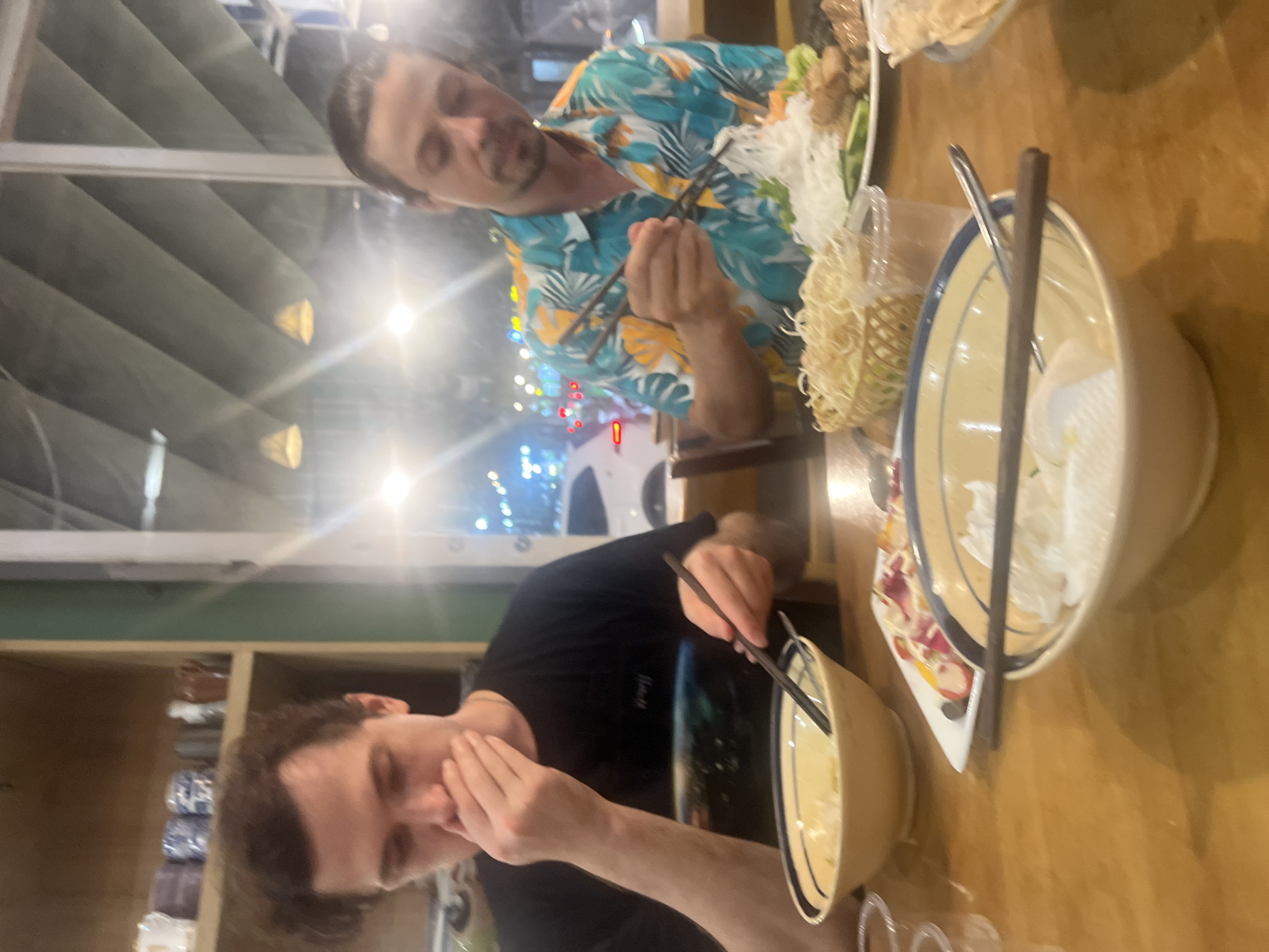 The image depicts two men seated at a wooden table in a restaurant, likely in Đà Nẵng, Vietnam, given the presence of Vietnamese cuisine and the distinctive lighting and window reflections suggesting an urban environment. The man on the left, wearing a black t-shirt, has his hand covering his mouth and is looking down at a bowl of food in front of him. He holds chopsticks. The man on the right, wearing a floral-patterned shirt, is actively eating with chopsticks, reaching for noodles from a plate containing meat, vegetables, and more noodles. Both bowls and plates appear to be part of a Vietnamese meal, possibly pho or bun cha. The restaurant interior is dimly lit, with the background showing reflections of city lights and what appears to be a window looking out onto a street. Shelving with ceramic containers is visible on the left side of the frame. The overall scene suggests a casual dining experience.
