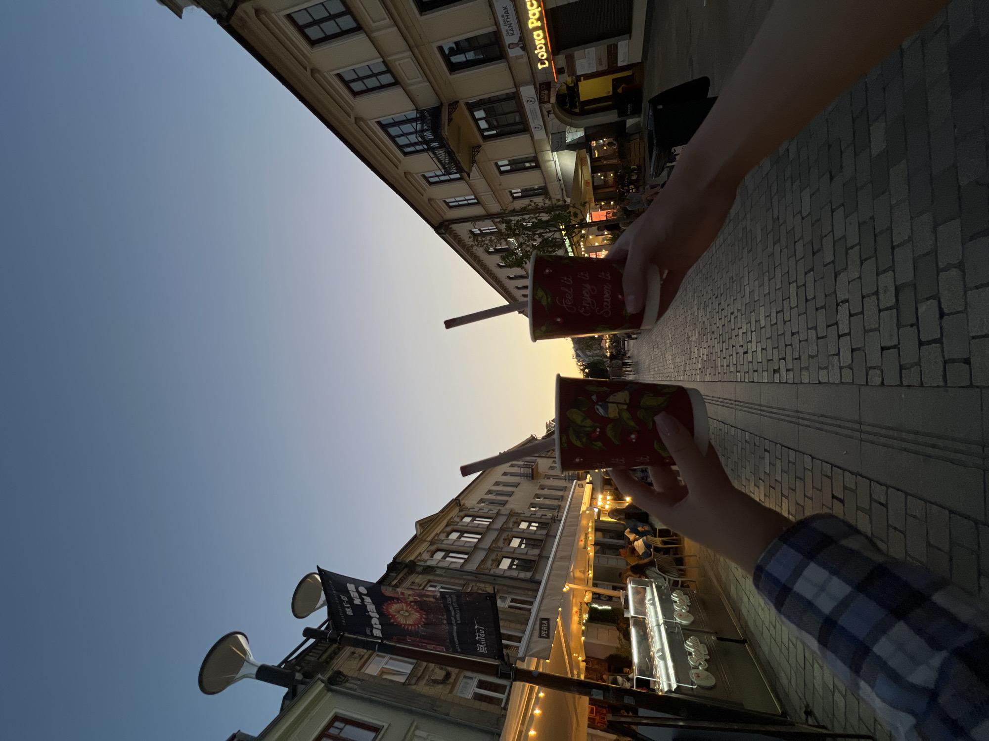 The image captures a street scene in the city of Lublin, Poland, during twilight. In the foreground, two hands are raised, each holding a red paper cup with a straw. The cups have a floral design and text that reads "Feel it Enjoy it Savor it". The hands are positioned in front of a cobblestone street lined with buildings and outdoor seating areas of cafes and restaurants. The sky is a clear gradient of blue and orange, indicating the sunset. On the left side of the street, a banner hangs from a lamppost, and illuminated signs for businesses are visible. People are seated at outdoor tables and walking along the street in the background, suggesting a lively evening atmosphere. The overall impression is one of leisure and enjoyment.